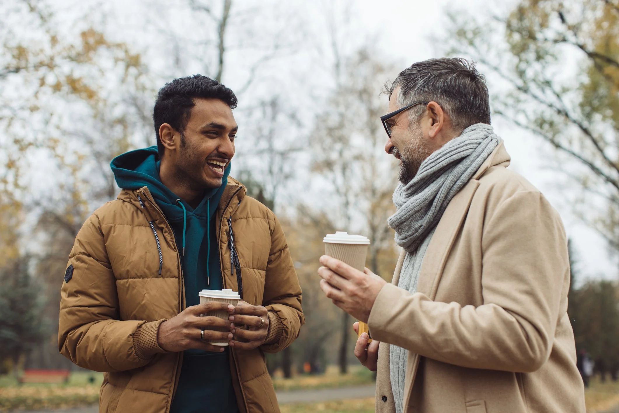 Two men enjoying a conversation outdoors, each holding a coffee cup, with trees and fall foliage in the background.