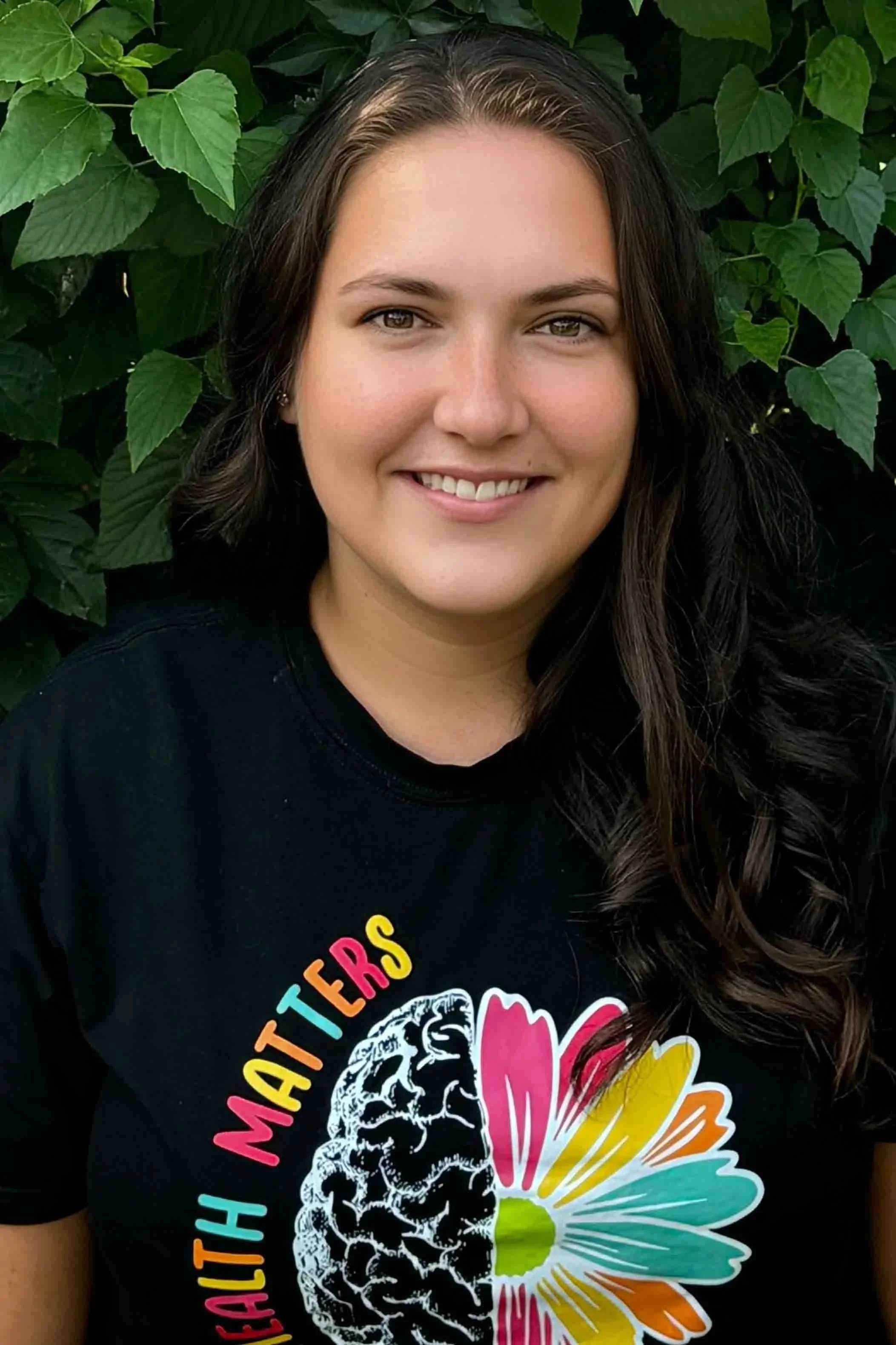 A smiling young woman with long wavy hair, wearing a black t-shirt with colorful graphics and text, standing in front of green leafy plants.