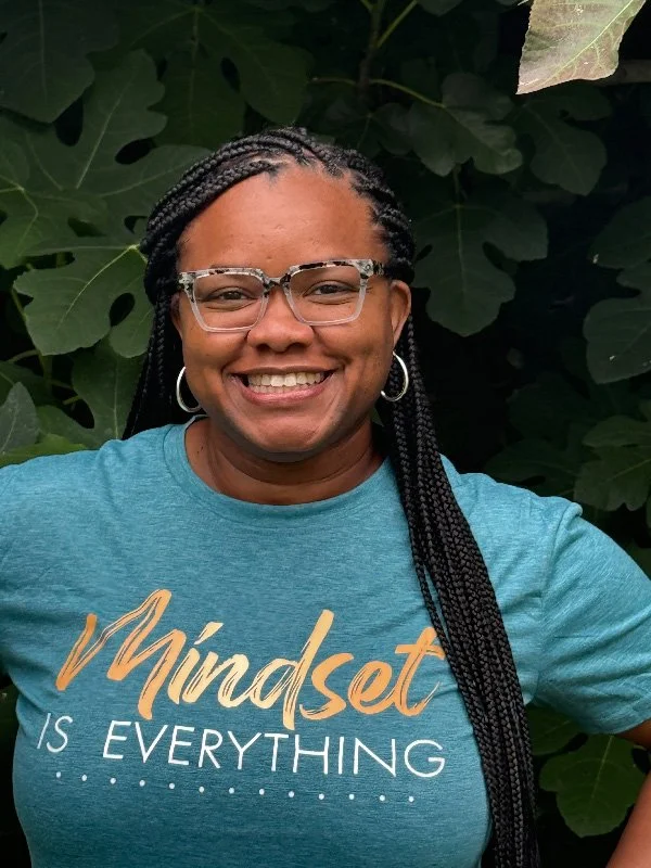 Female Therapist in Fredericksburg Virginia that does telehealth  smiling, wearing glasses and a teal shirt with the text 'Mindset is Everything,' standing in front of leafy greenery.