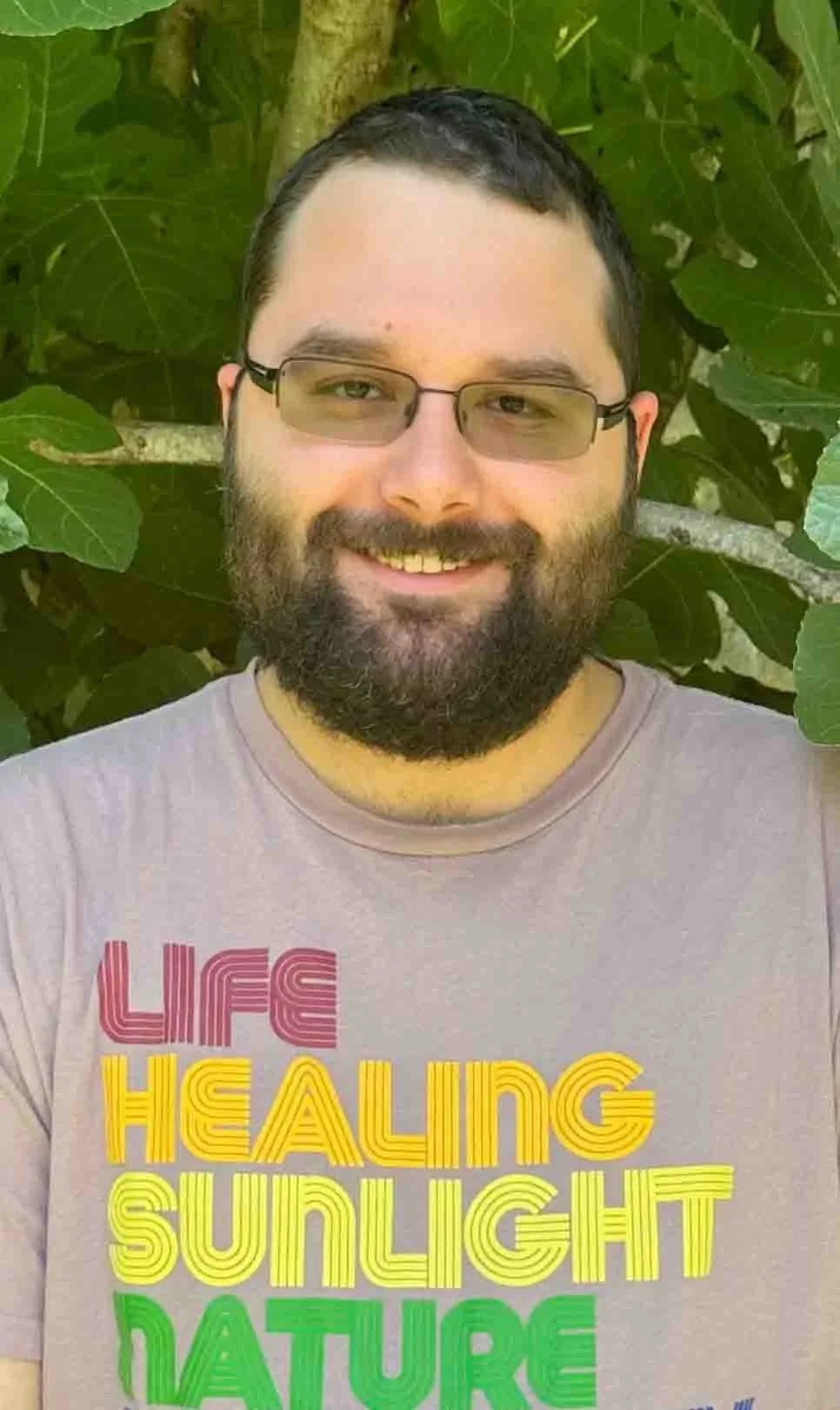 A smiling man with glasses and a beard standing outdoors in front of green foliage, wearing a t-shirt with multicolored text that reads, 'Life Healing Sunlight Nature'.