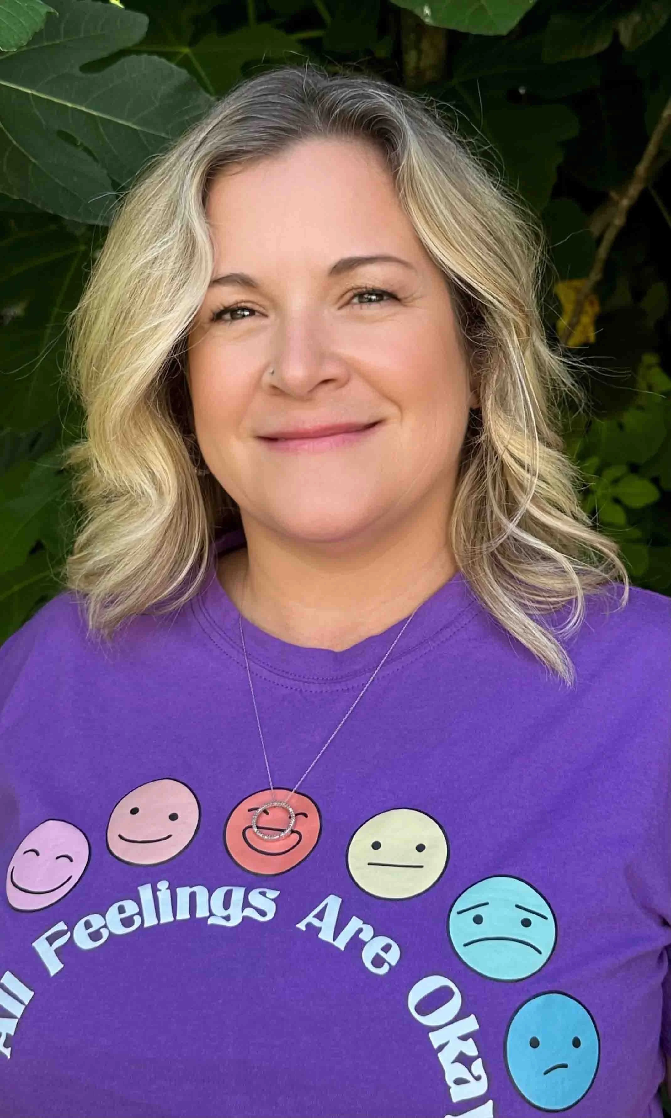 A woman with blonde curl hair wearing a purple shirt with various emoji faces and the text 'All Feelings Are O' standing outdoors in front of green leaves.