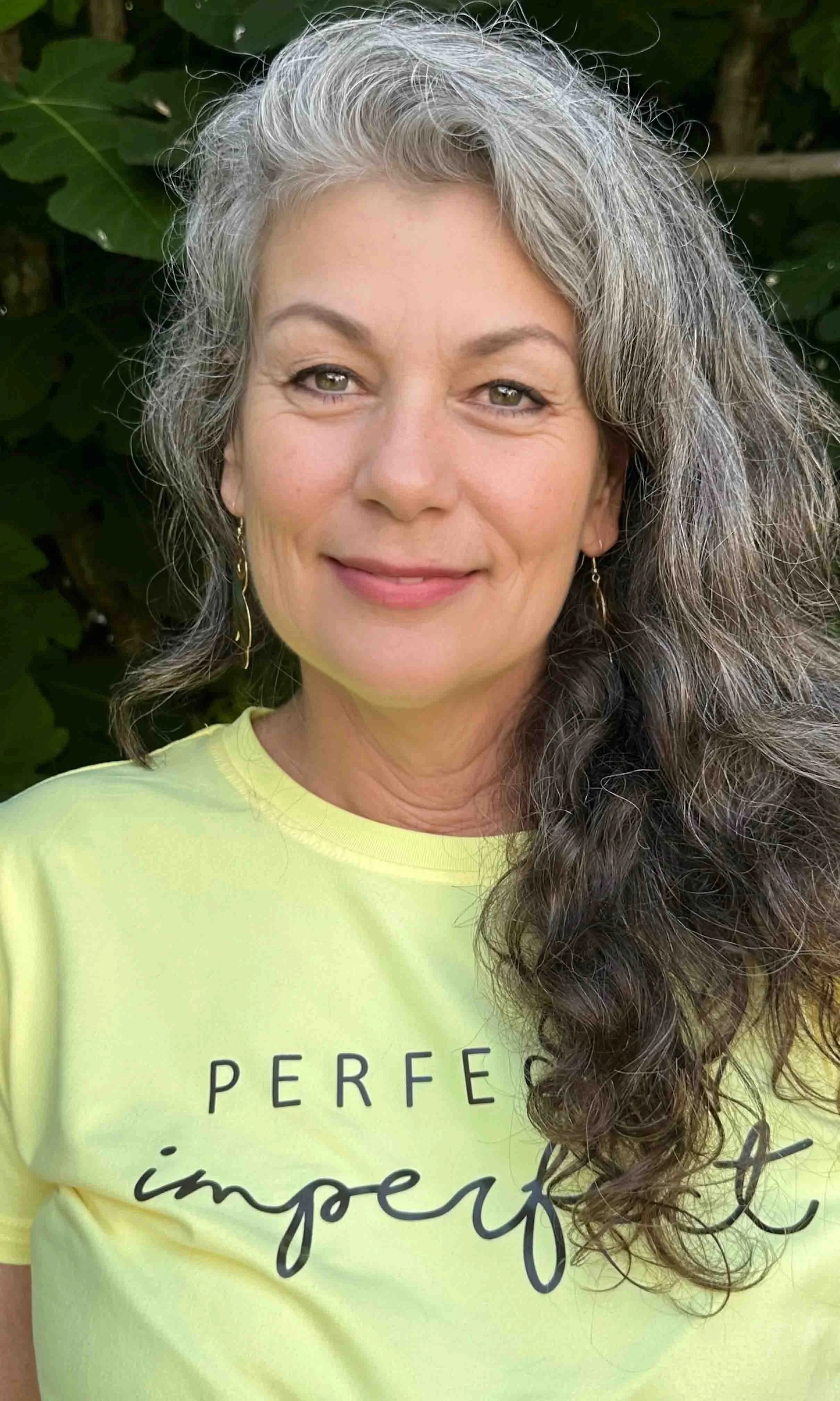 A smiling woman with gray and brown hair, wearing a yellow t-shirt with the words 'PERFECT imperfect' written on it, standing outdoors in front of green foliage.