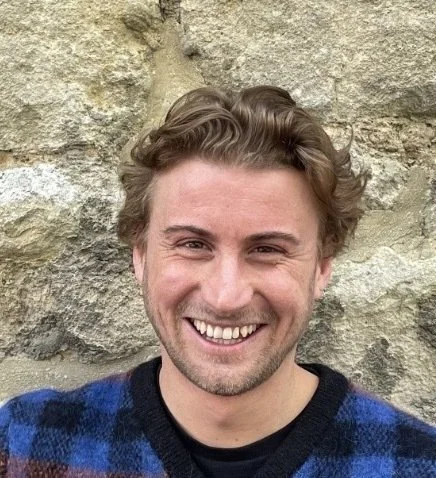 A smiling man with wavy hair and facial hair, standing in front of a stone wall, wearing a dark sweater with a plaid pattern.