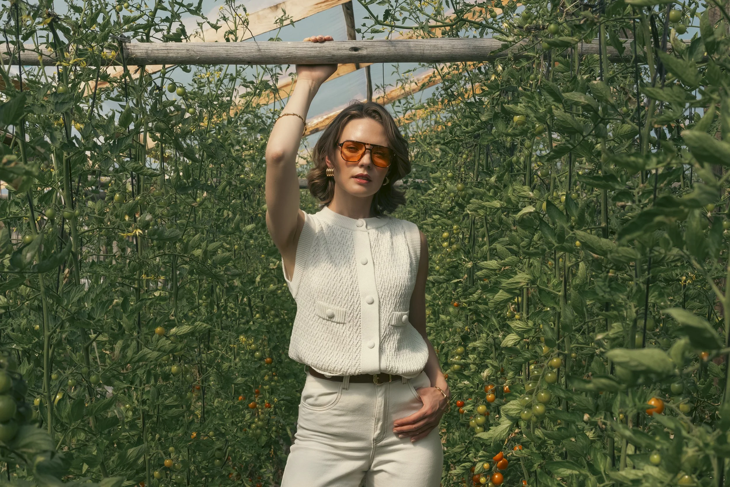 Una mujer con gafas anaranjadas, vestida con una blusa blanca y pantalones beige, posa en un invernadero de tomates verdes y maduros.