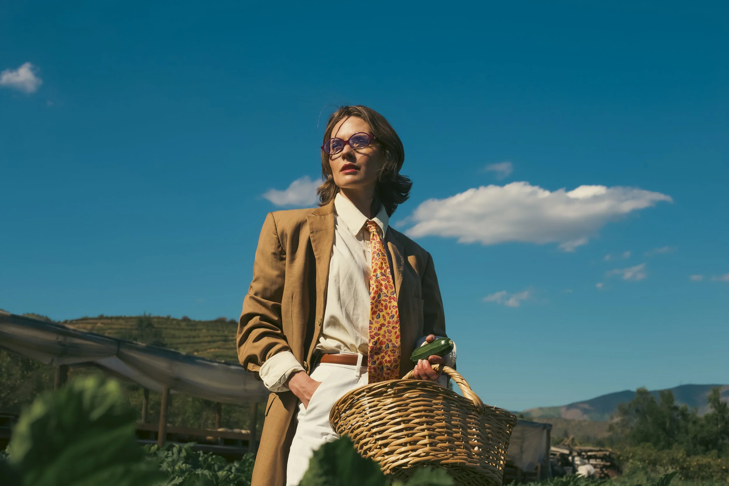 Mujer con gafas de sol y ropa vintage sosteniendo una cesta en un campo con montañas al fondo
