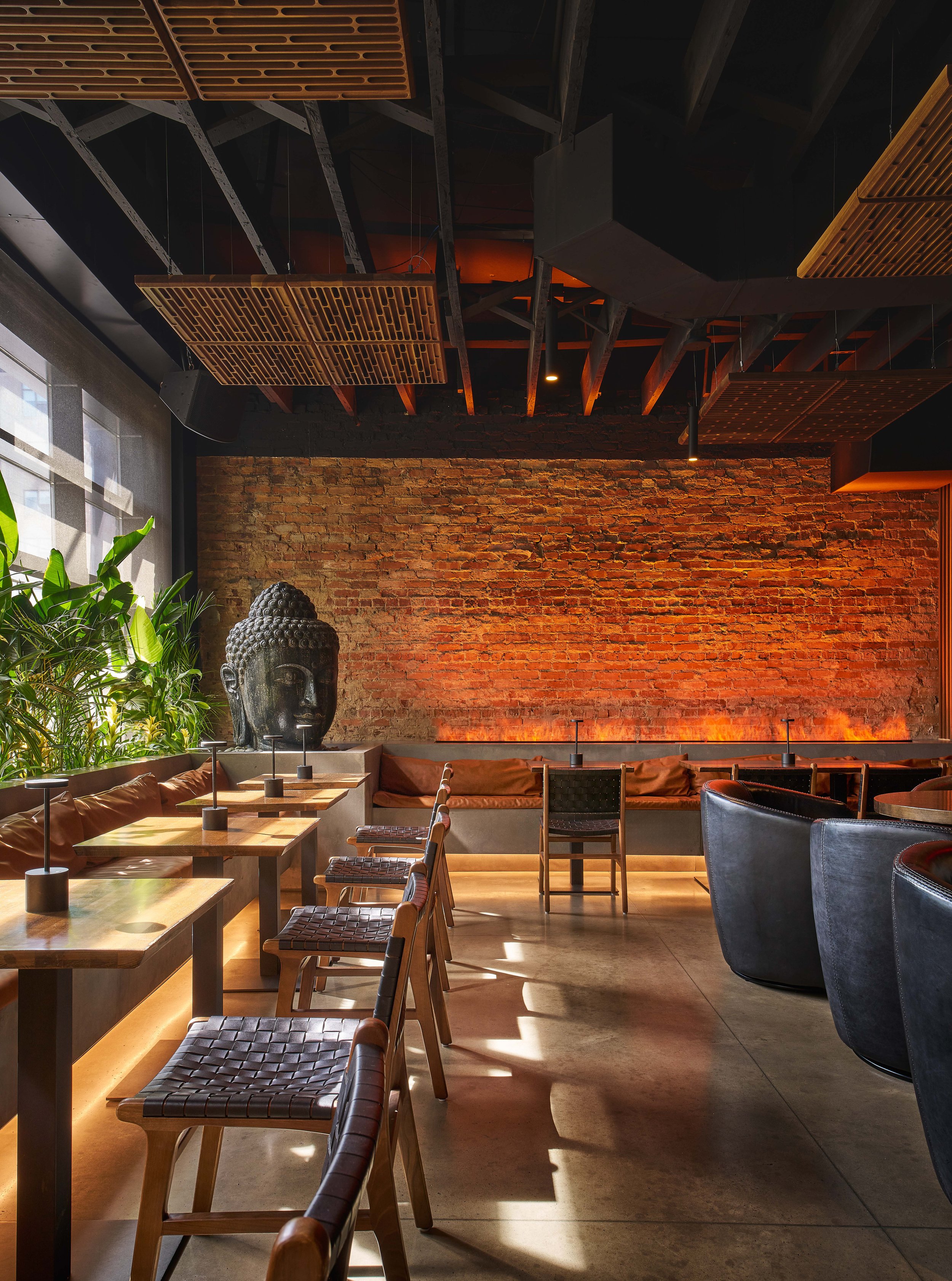 Interior of a modern Tulum, Mexico and Bali-inspired listening lounge, cocktail bar, sound bar, restaurant or cafe with tables, leather chairs, a brick wall and organic environment. There are real plants, a Buddha head fountain, and large windows.