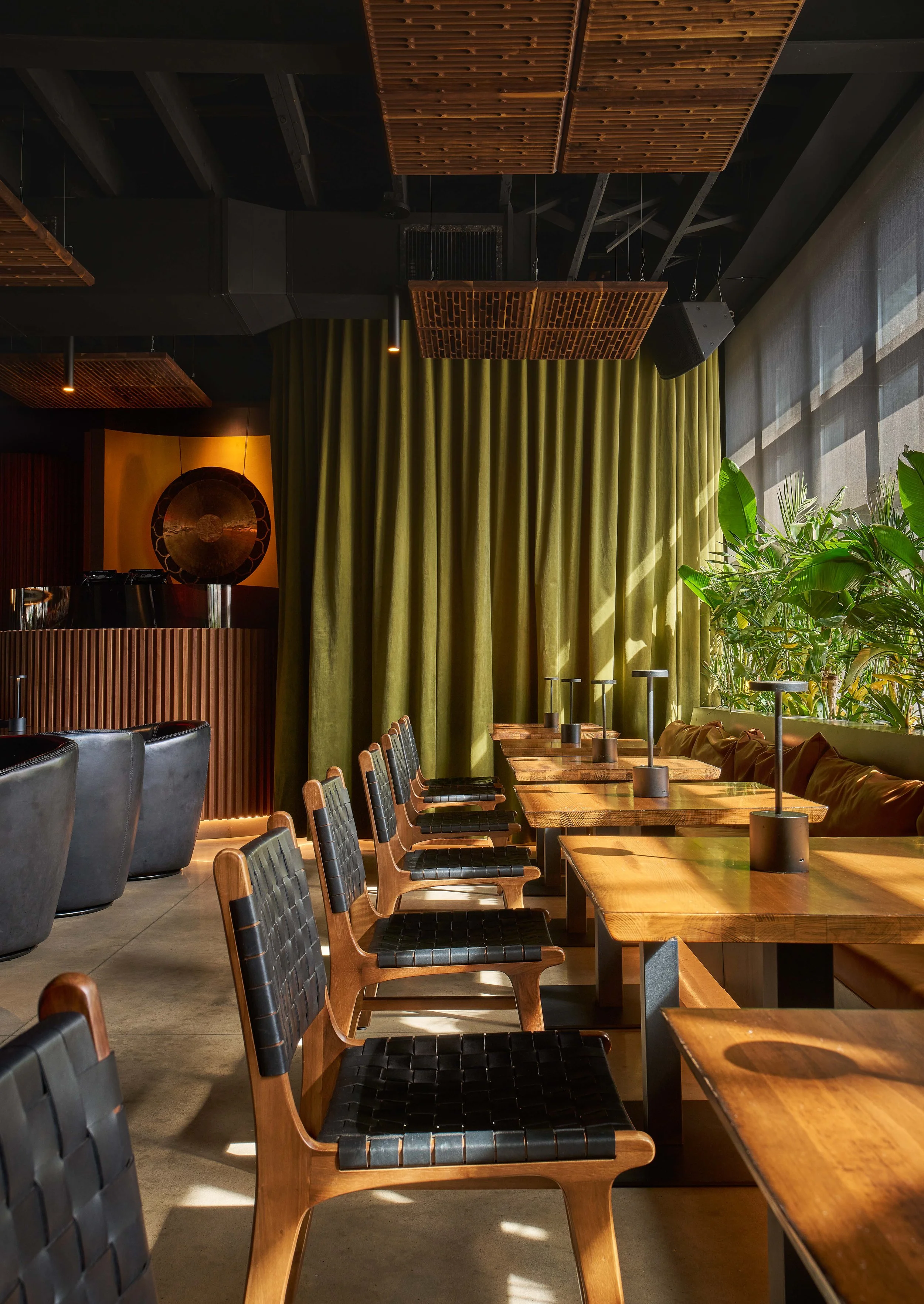 Interior of a modern restaurant, cocktail bar or cafe with wooden tables, black leather chairs, lush green plants, velvet curtains, decorative ceiling panels, a chandelier and leather booths.
