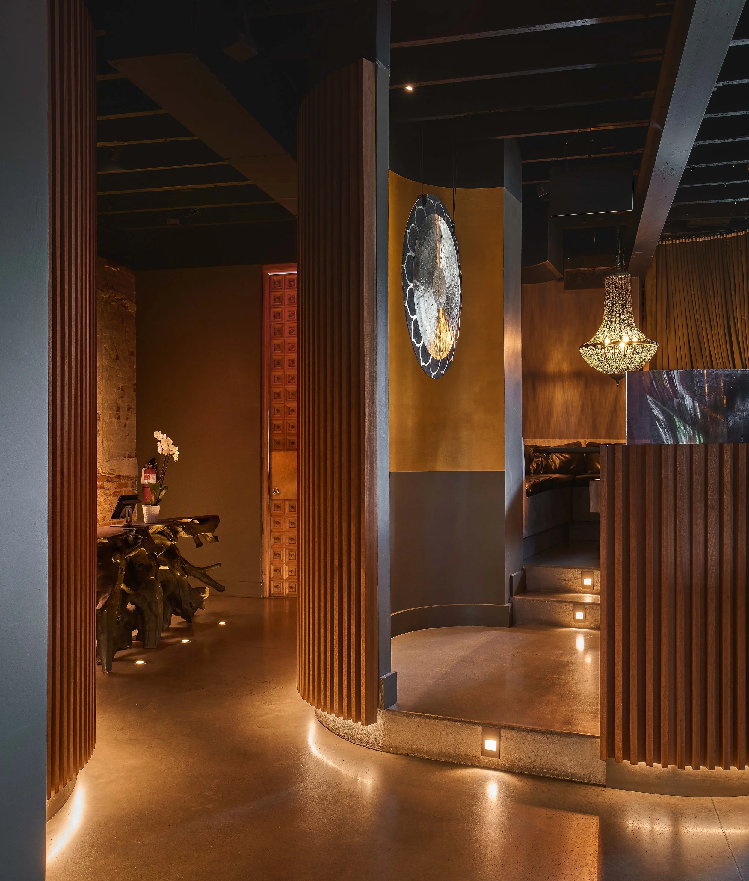 Interior view of a modern space with mahogany wood slat walls, warm lighting, a large decorative sculpture, and a chandelier. There is a curved wall with steps and small recessed lights.