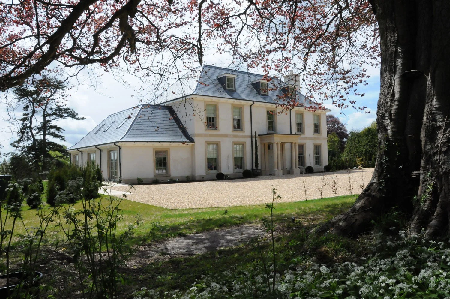 Alex Oliver Associates Ltd — A large, elegant two-story house with a slate roof and white facade, surrounded by trees and greenery, viewed from a garden area with a large foregrou