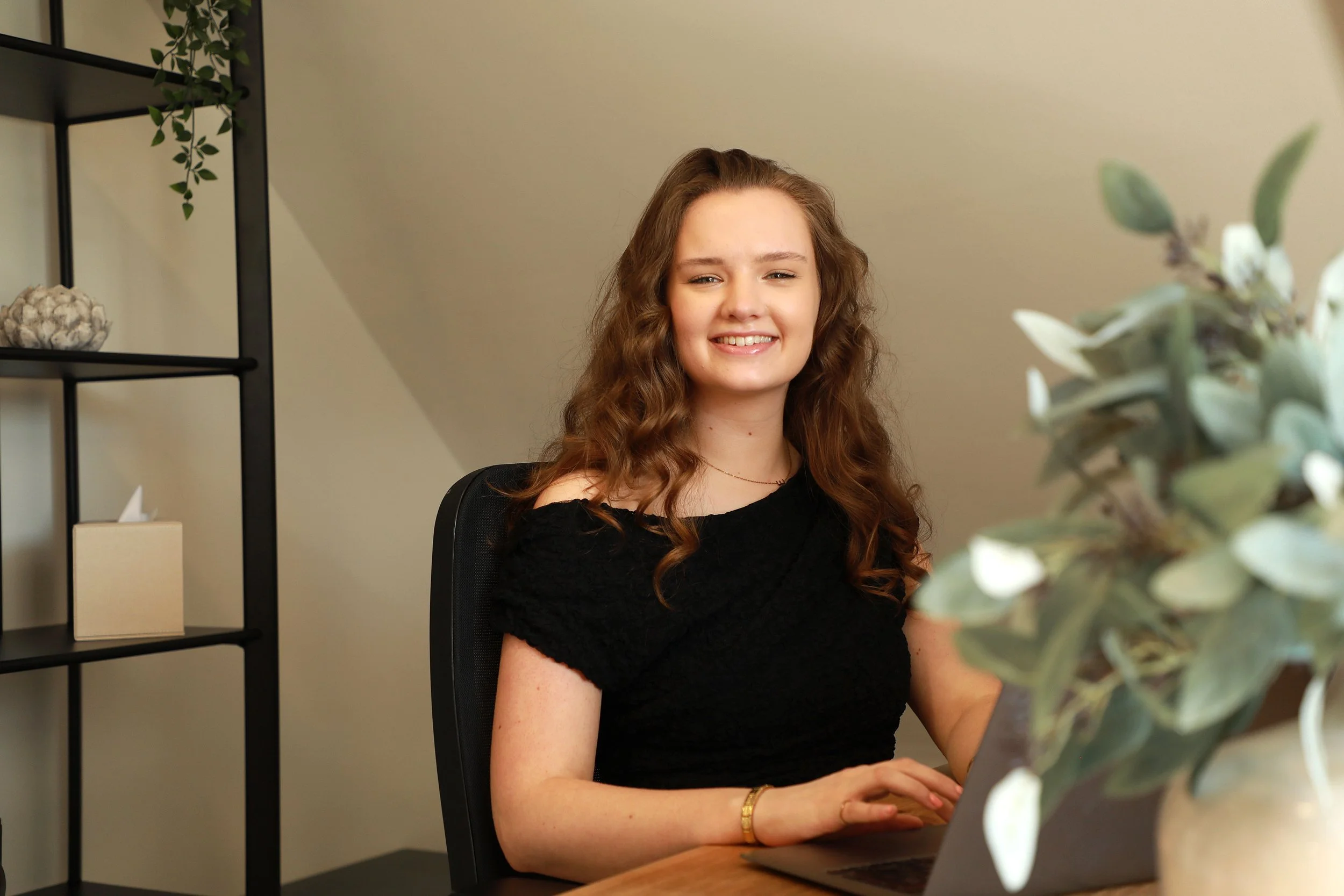A young woman with long, curly brown hair smiling while sitting at a desk with a laptop, in a room decorated with a black metal shelving unit with decorative items, and a potted plant with green leaves in the foreground.