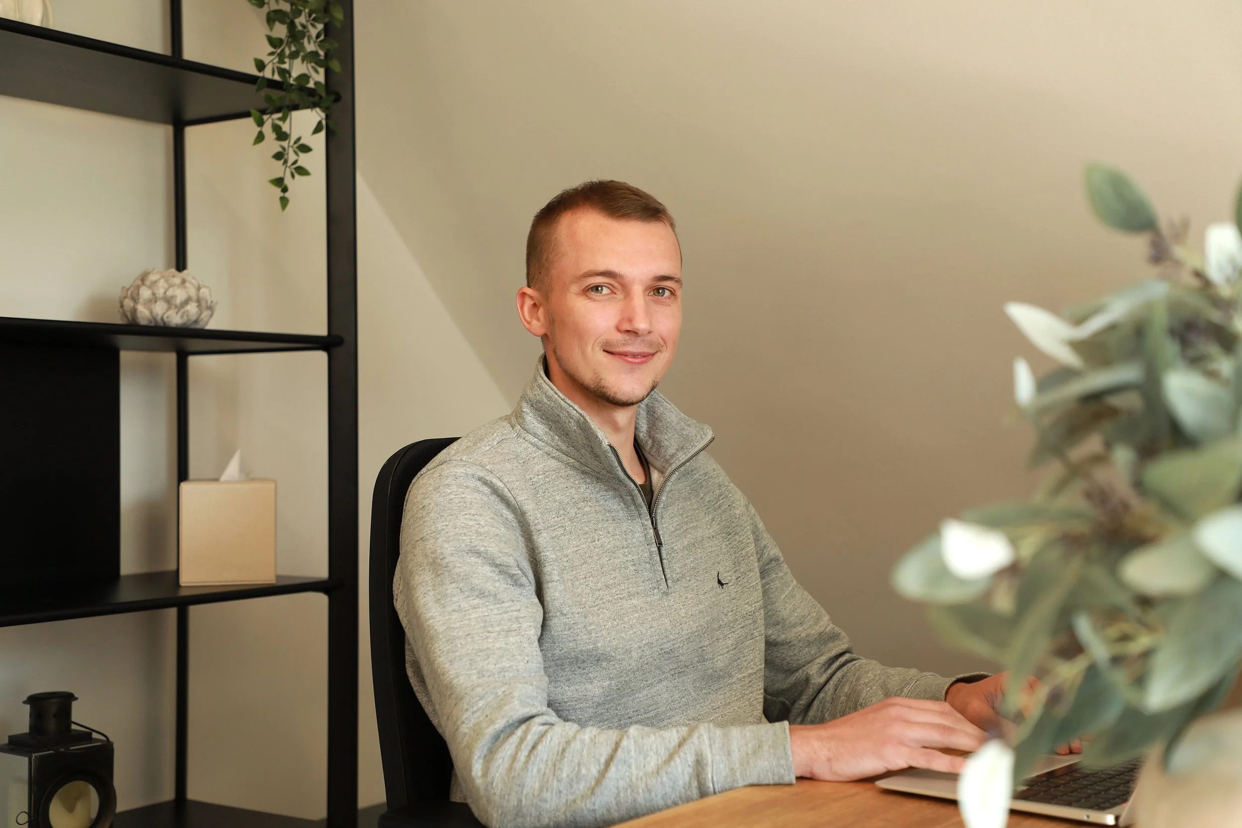 MatchingUp Staffing. A young man with short light brown hair and fair skin, sitting at a desk with a laptop, smiling at the camera. He is wearing a light gray quarter-zip sweater.