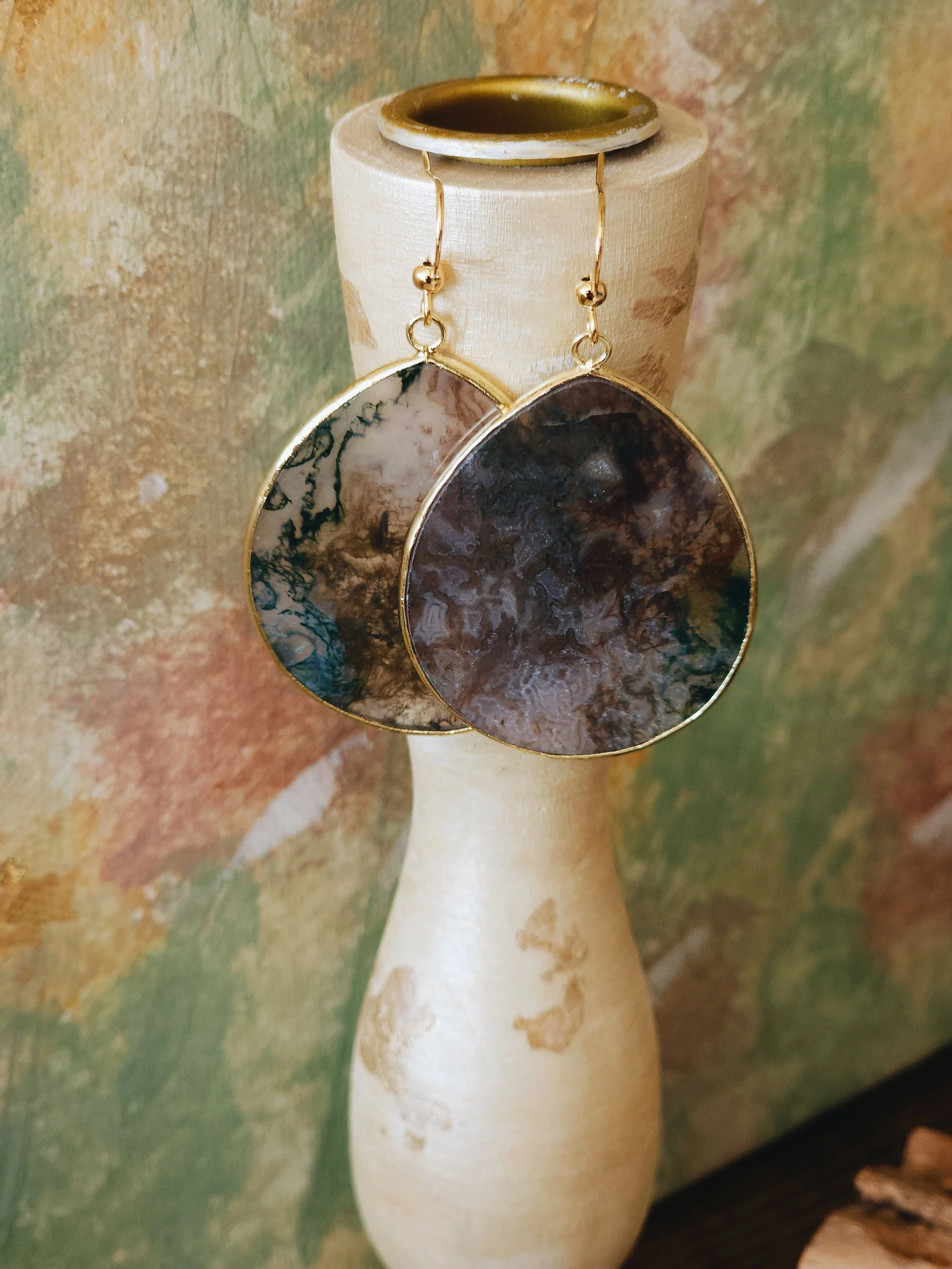 Pair of large teardrop-shaped earrings with marbled black, brown, and cream stones, hanging from a light-colored vase against a textured, multicolored background.