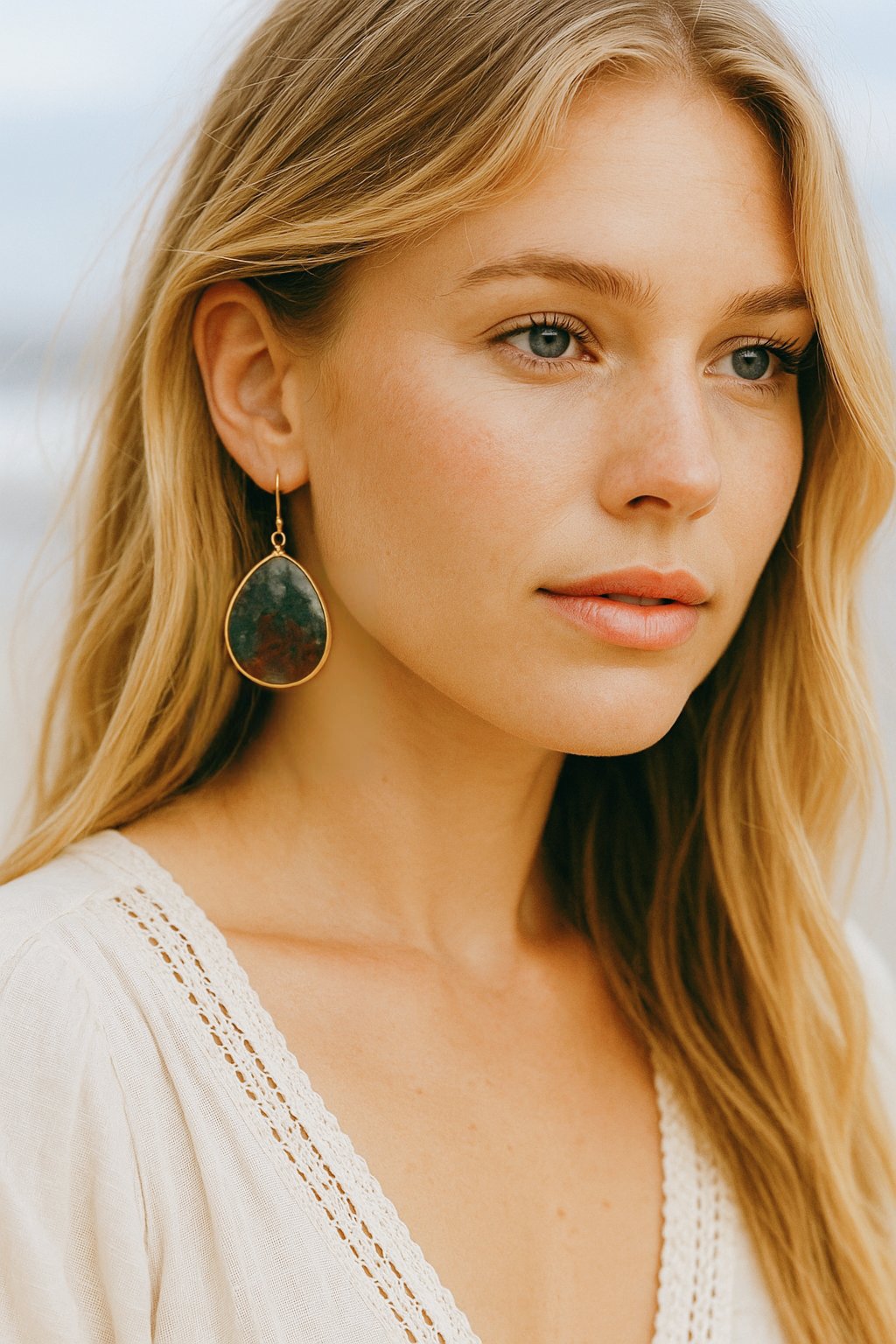 A close-up portrait of a young woman with long blonde hair, blue eyes, wearing a white top with lace detail, and a large teardrop-shaped earring with a dark-colored stone.
