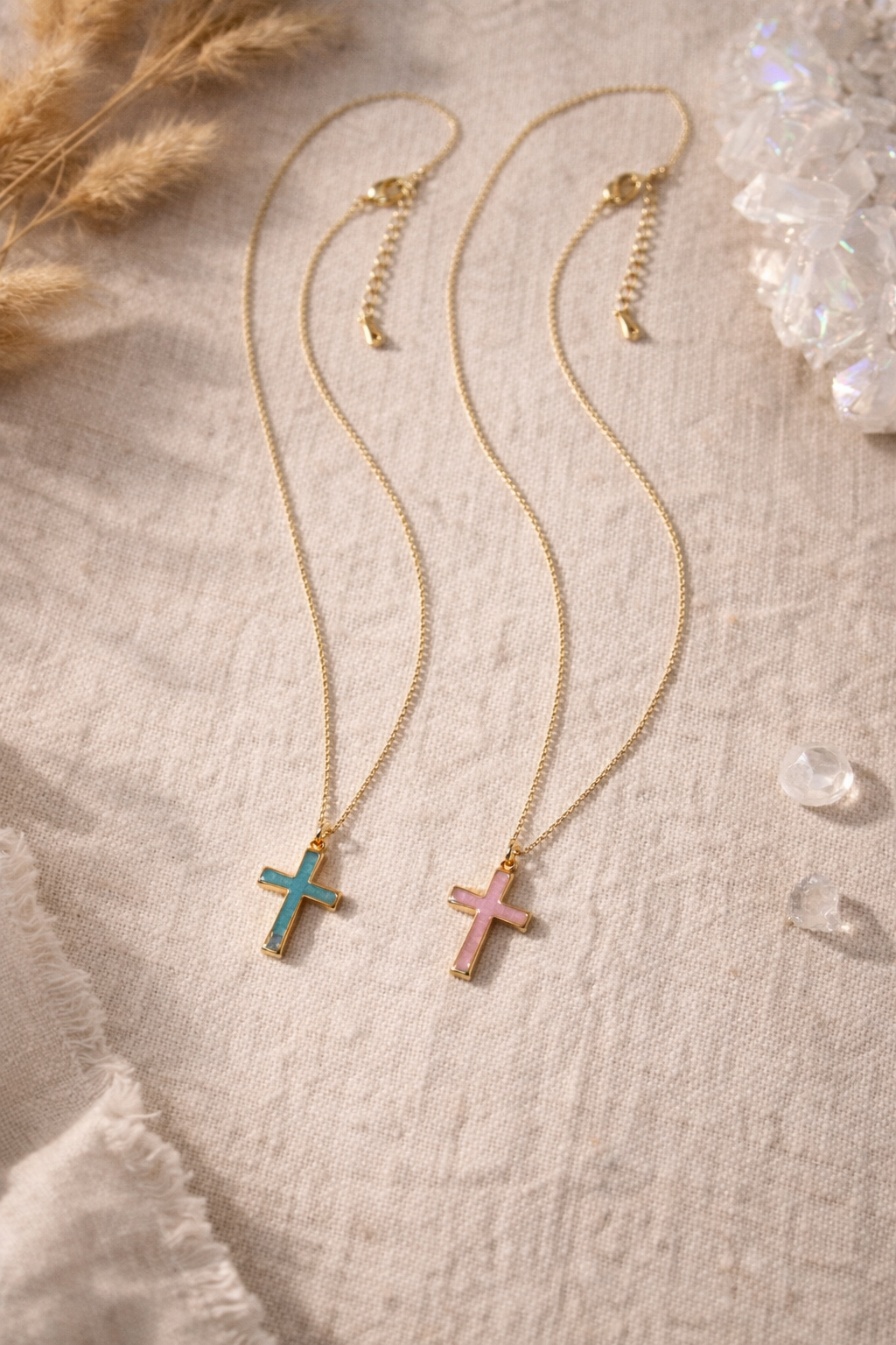 Two gold chain necklaces with cross pendants, one with a blue cross and the other with a pink cross, laid on a beige fabric surface with decorative dried flowers and clear beads.