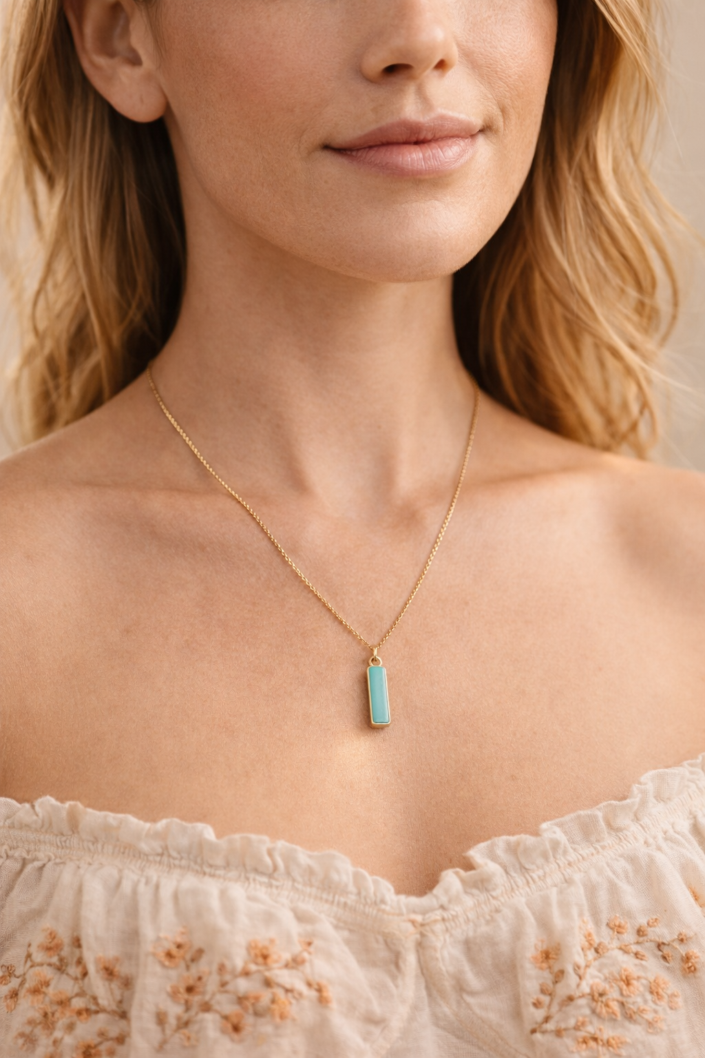 A woman with fair skin and red hair wearing a gold chain necklace with a rectangular light blue pendant, dressed in an off-shoulder, cream-colored top with orange embroidery.