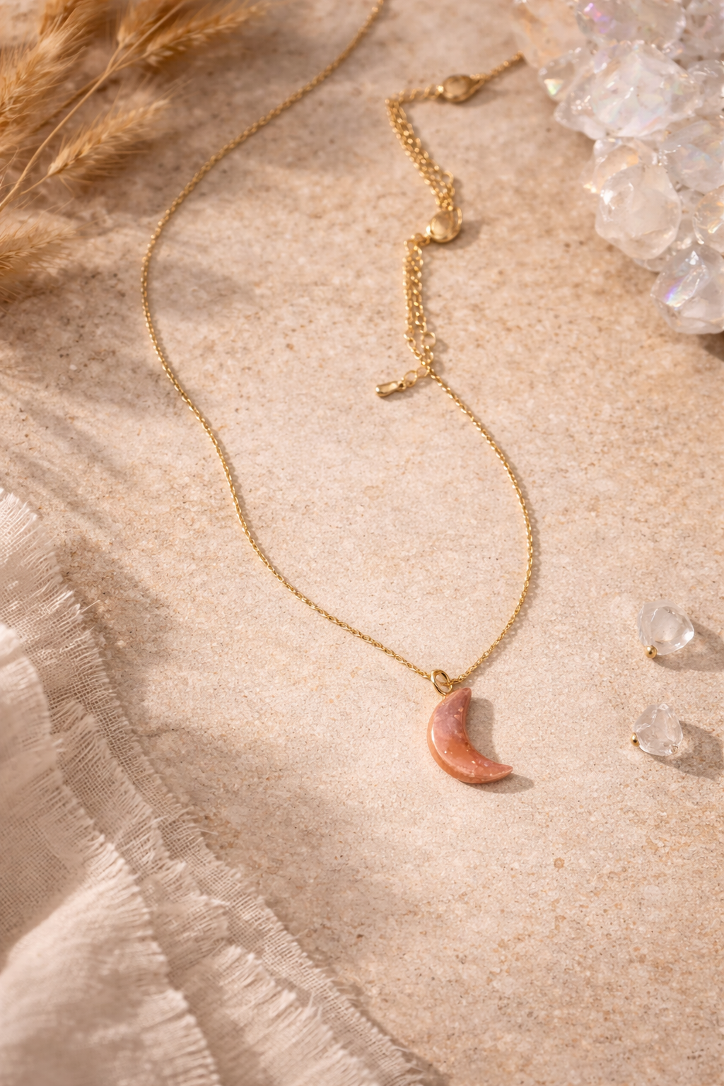 Gold necklace with a pink moon-shaped pendant on a sandy surface, surrounded by small clear gemstones, some dried plants, and fabric.