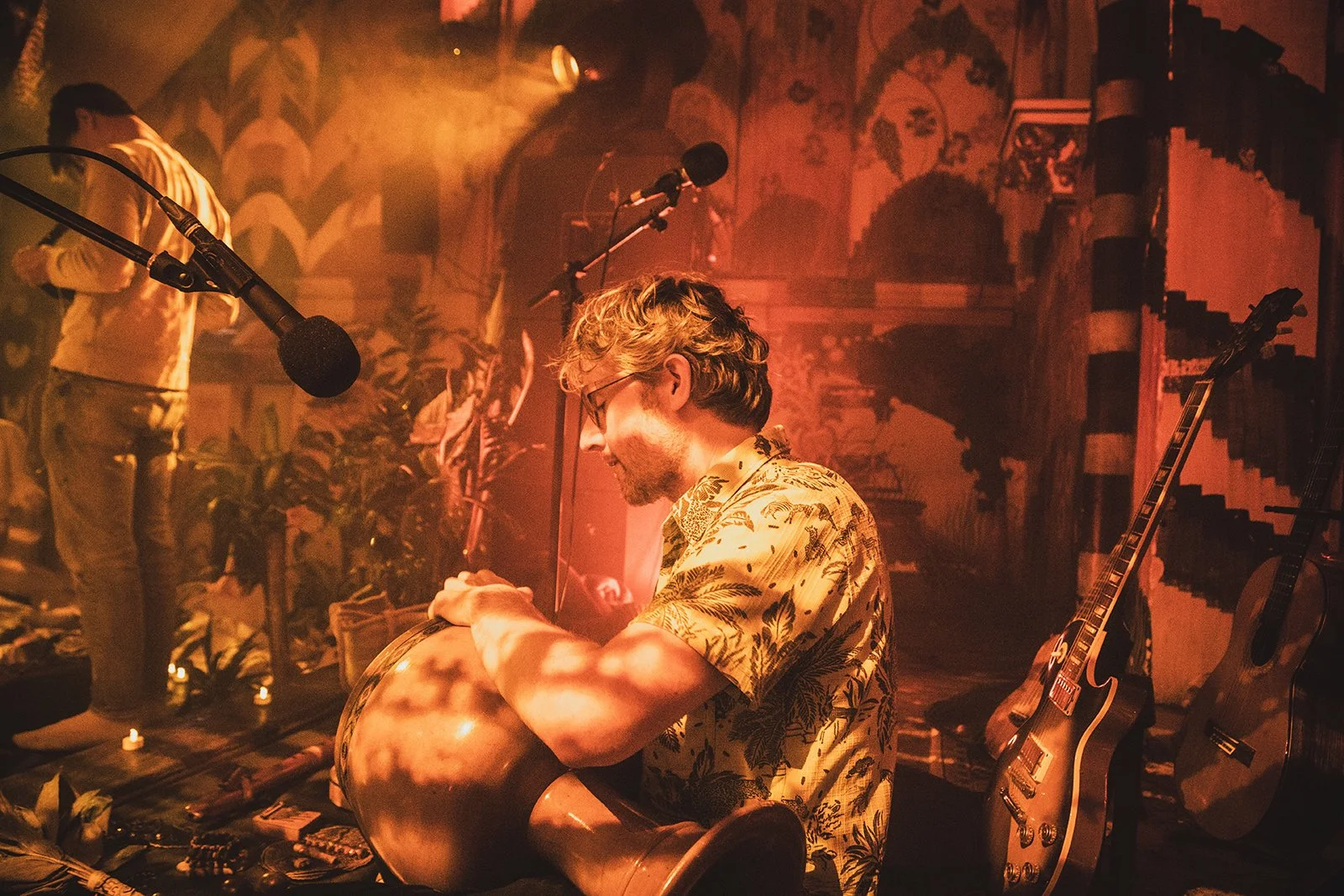A musician with curly hair and glasses is sitting and playing a djembe drum on stage, surrounded by guitars, with a performer in the background and stage lighting creating a warm, atmospheric ambiance.