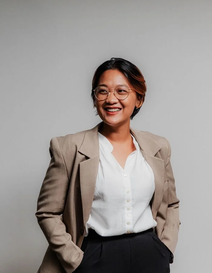 A smiling woman with short dark hair, wearing glasses, a beige blazer, white blouse, and black pants, posing against a plain gray wall.