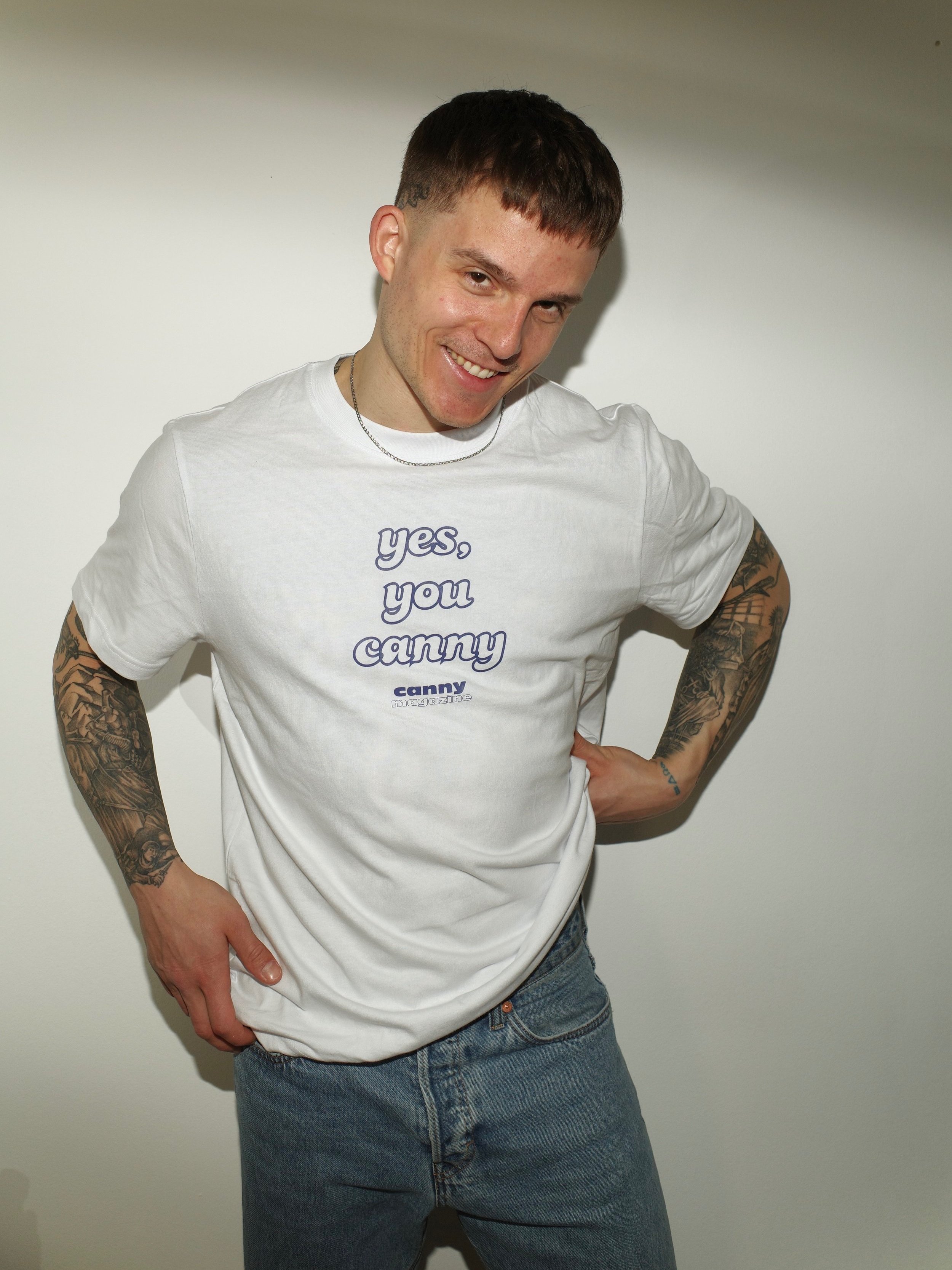 canny Merch: Weißes T-Shirt mit blauer Aufschrift an einem Mann mit Tattoos.
