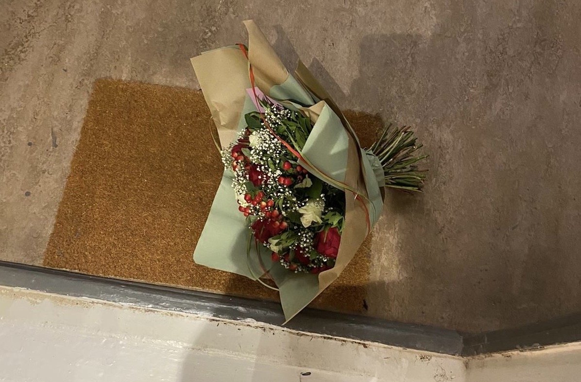 Auf der Fußmatte vor einer Wohnungstür liegt ein Blumenstrauß. Er ist aus roten und weißen Rosen mit Hagebutten verziert.