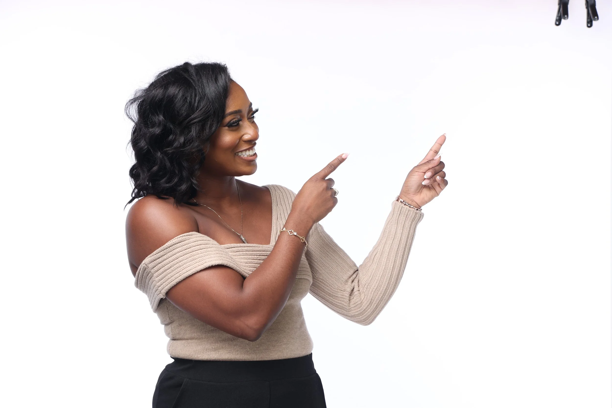 A woman smiling and pointing to her right, wearing a beige off-shoulder sweater and black pants, with a plain white background.