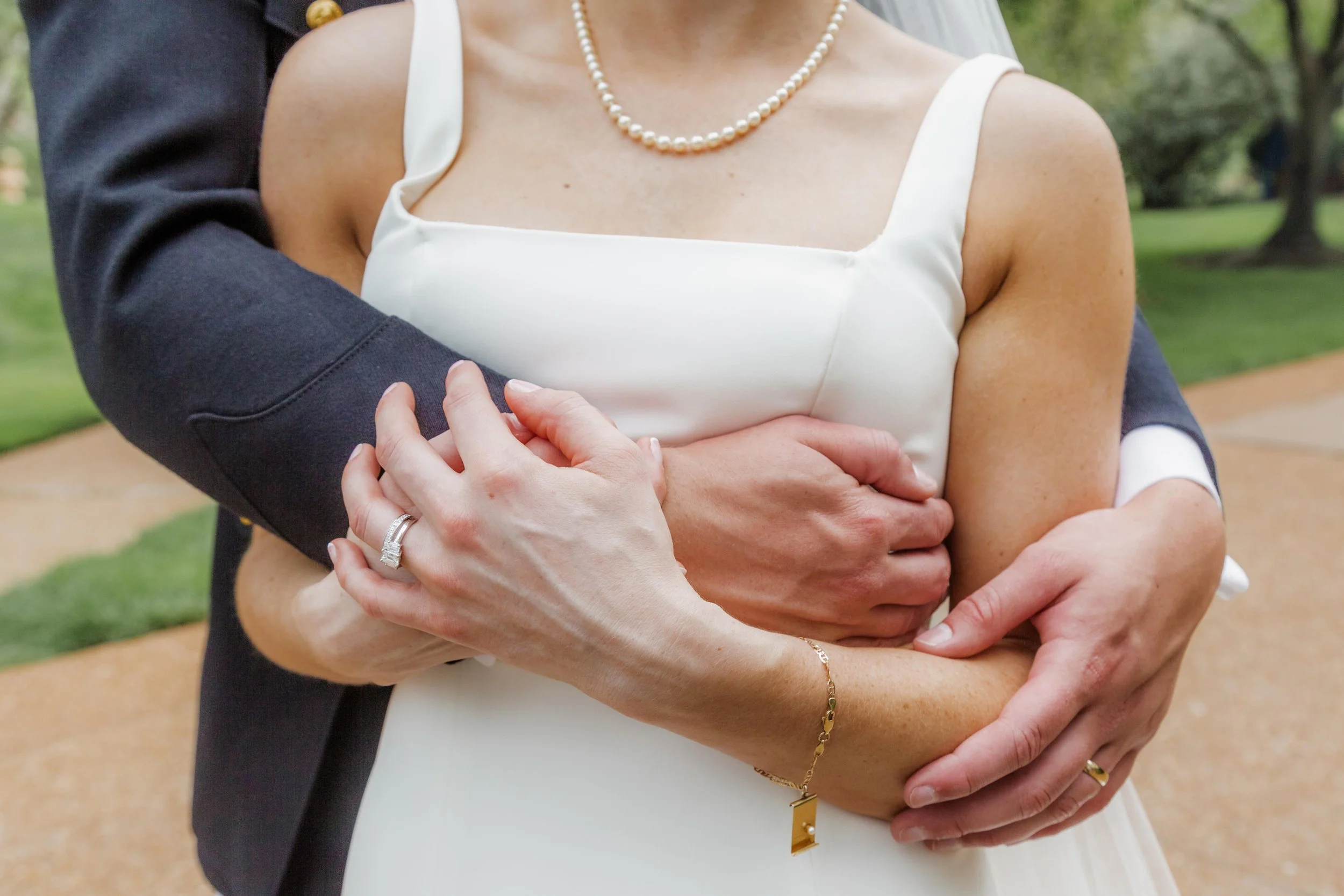 A couple dressed in wedding attire, with the groom embracing the bride from behind, their arms intertwined, outdoors in a park-like setting.