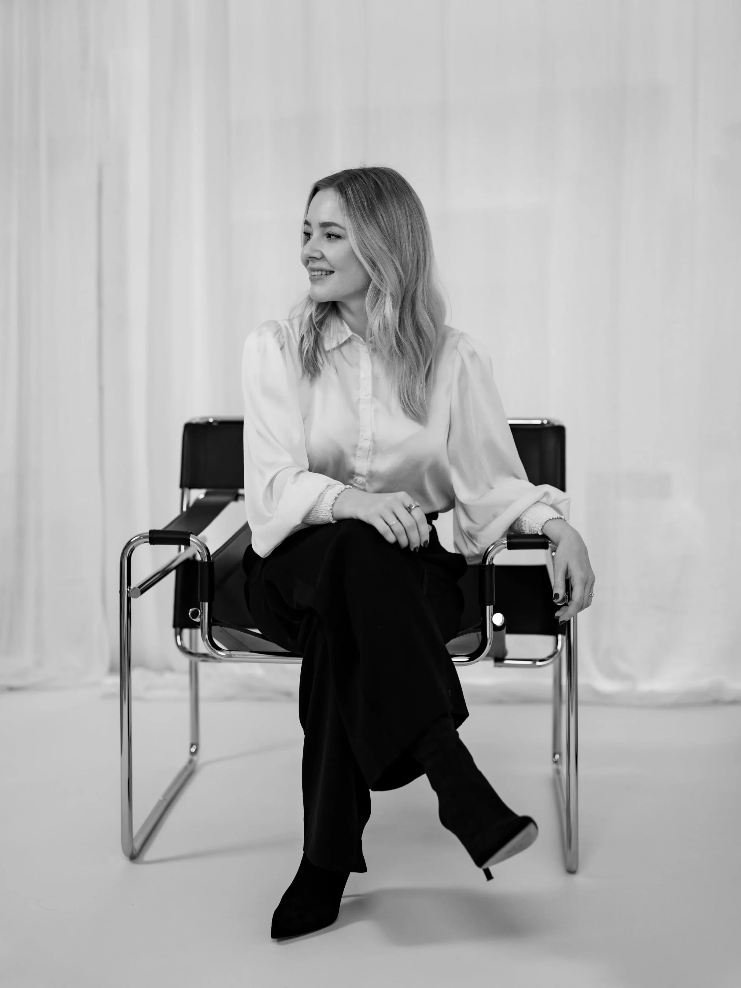 Schwarz-weiß-Foto einer Frau, die auf einem Stuhl sitzt, lächelt und nach links schaut, mit lockigem, schulterlangem Haar, weißem Hemd, schwarzen Hosen und Stiefeln.