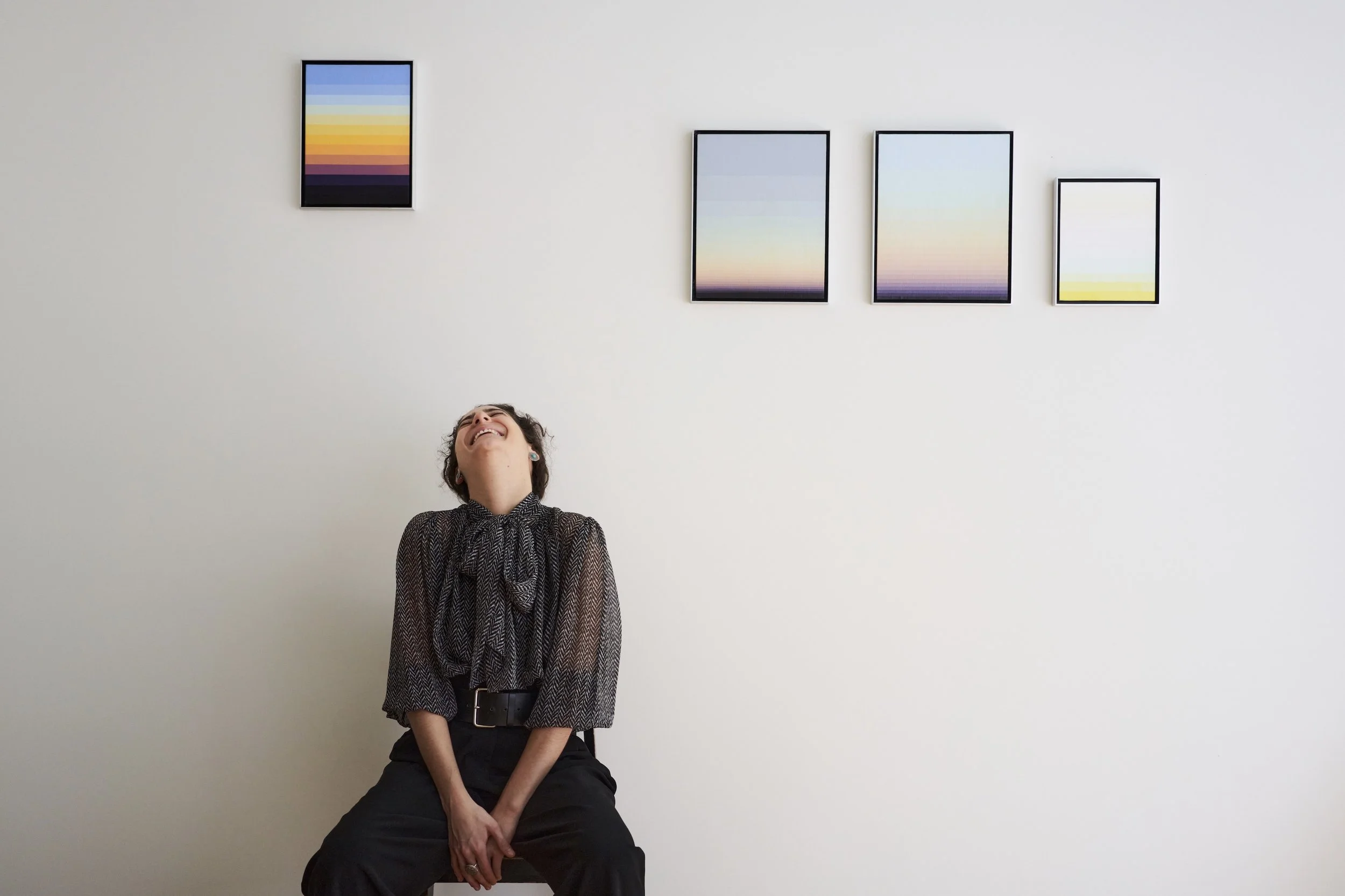 A woman with dark hair smiling and looking upward, sitting against a white wall with four framed art pieces depicting colorful abstract gradients.