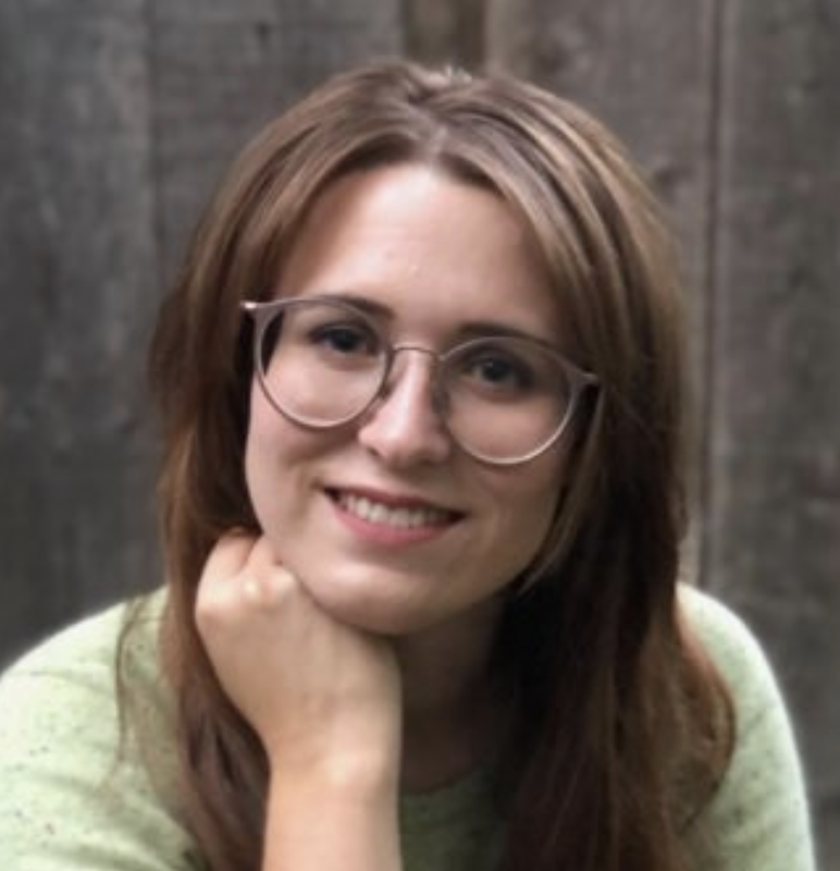 A young woman with shoulder-length brown hair and glasses, smiling and resting her chin on her hand in front of a wooden background.