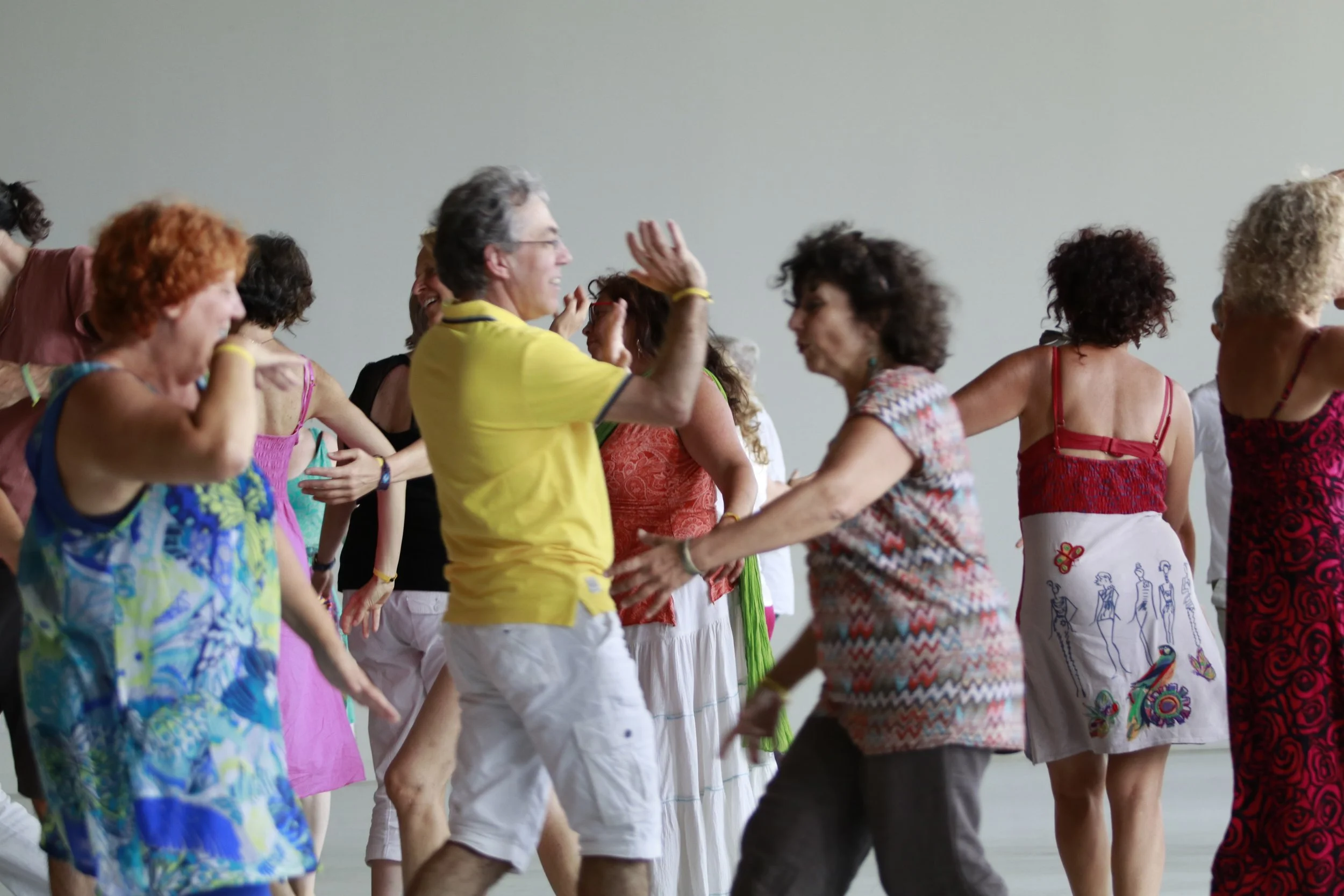 Group of adults dancing together indoors, smiling, wearing colorful casual summer clothes.