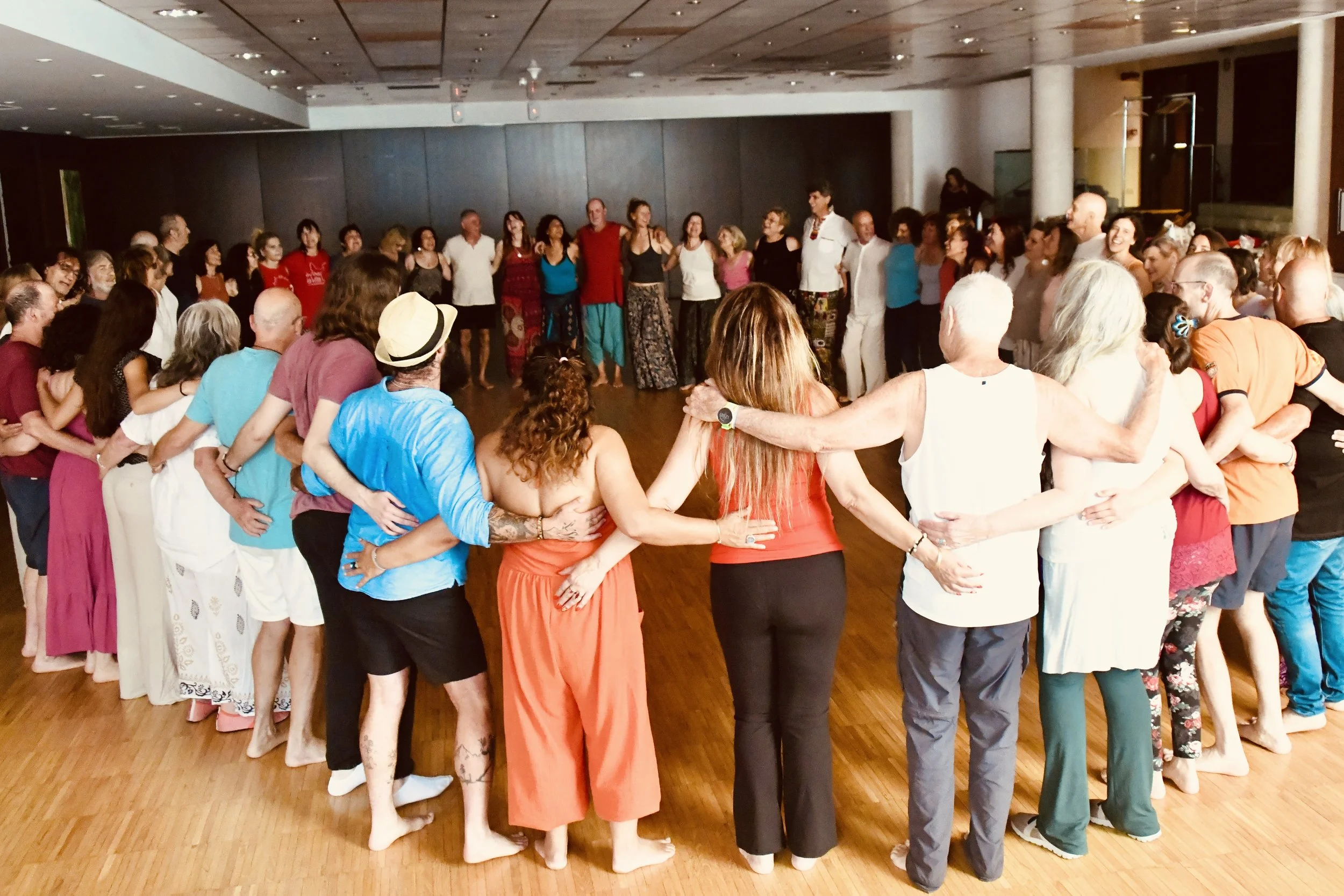 A diverse group of people holding hands in a circle, participating in a dance or celebration on a wooden floor in a spacious indoor venue.