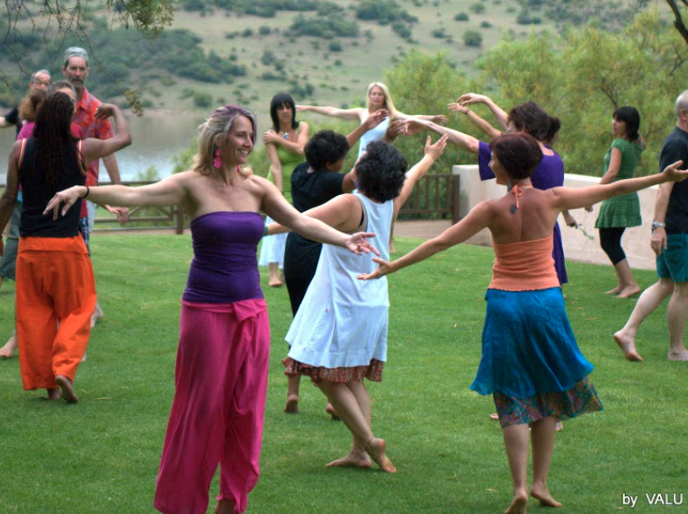 People dancing outdoors on grass with arms outstretched, colorful clothing, lush trees, and a lake in the background.