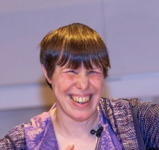 Person with short brown hair smiling widely, showing teeth, wearing a purple top and a checkered jacket. CArol Pound, Kavina, Biodanza Greenwich london