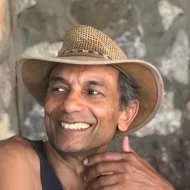 Smiling man wearing a wide-brimmed straw hat and a dark tank top, outdoors with a stone wall in the background. Altair de Almeida, biodanza malvern