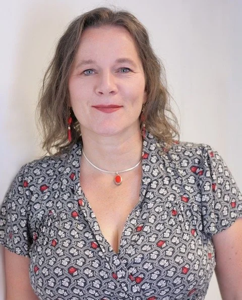 A woman with wavy, shoulder-length hair smiling at the camera, wearing a patterned blouse with red and black accents, a silver necklace with an orange pendant, and matching earrings. Ali baker, biodanza lancashire, yorkshire, lancaster