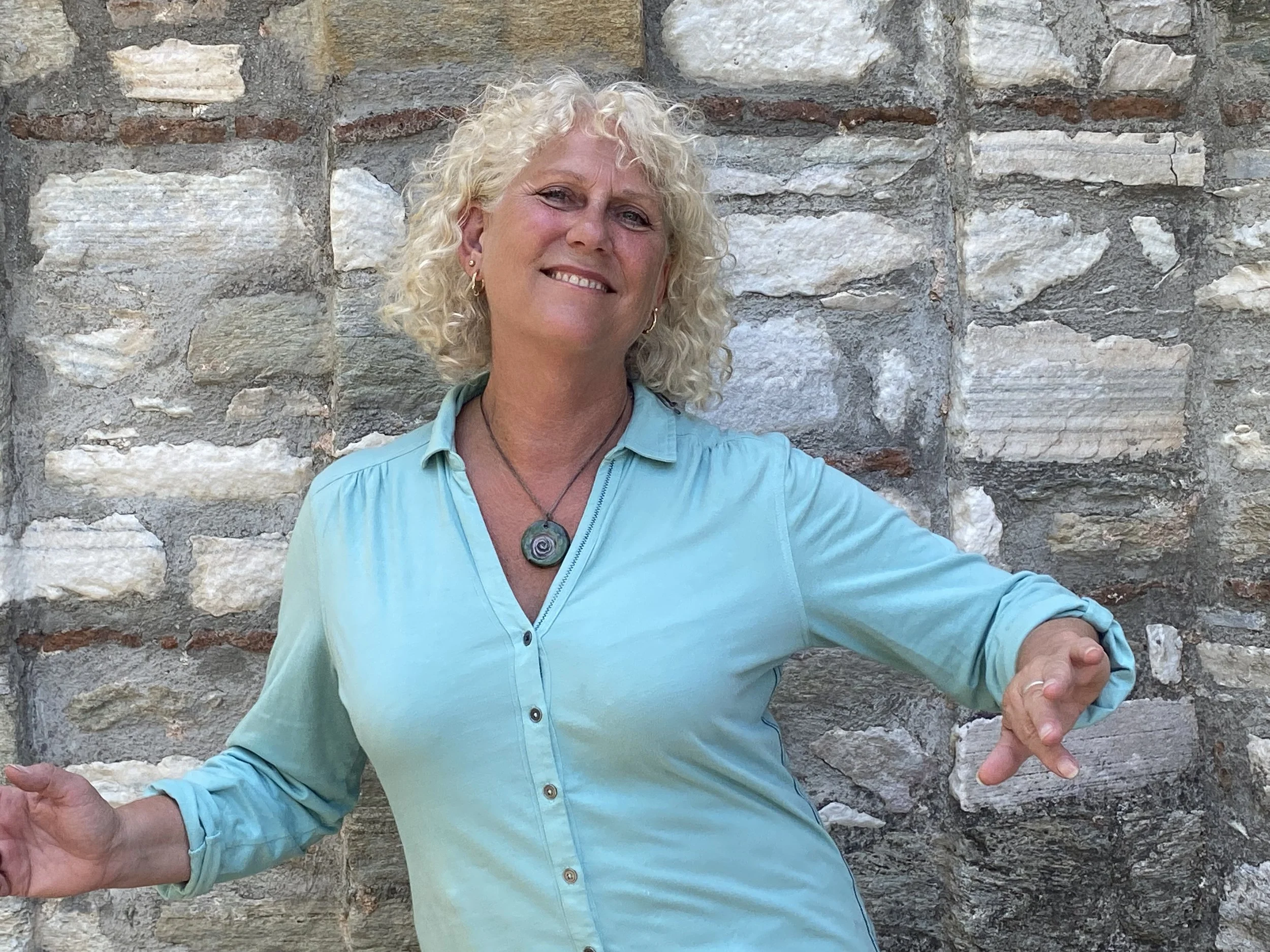 A smiling woman with curly blonde hair, dressed in a light blue button-up shirt with rolled sleeves, standing in front of a stone wall. Tracy Seed, Biodanza london and greece