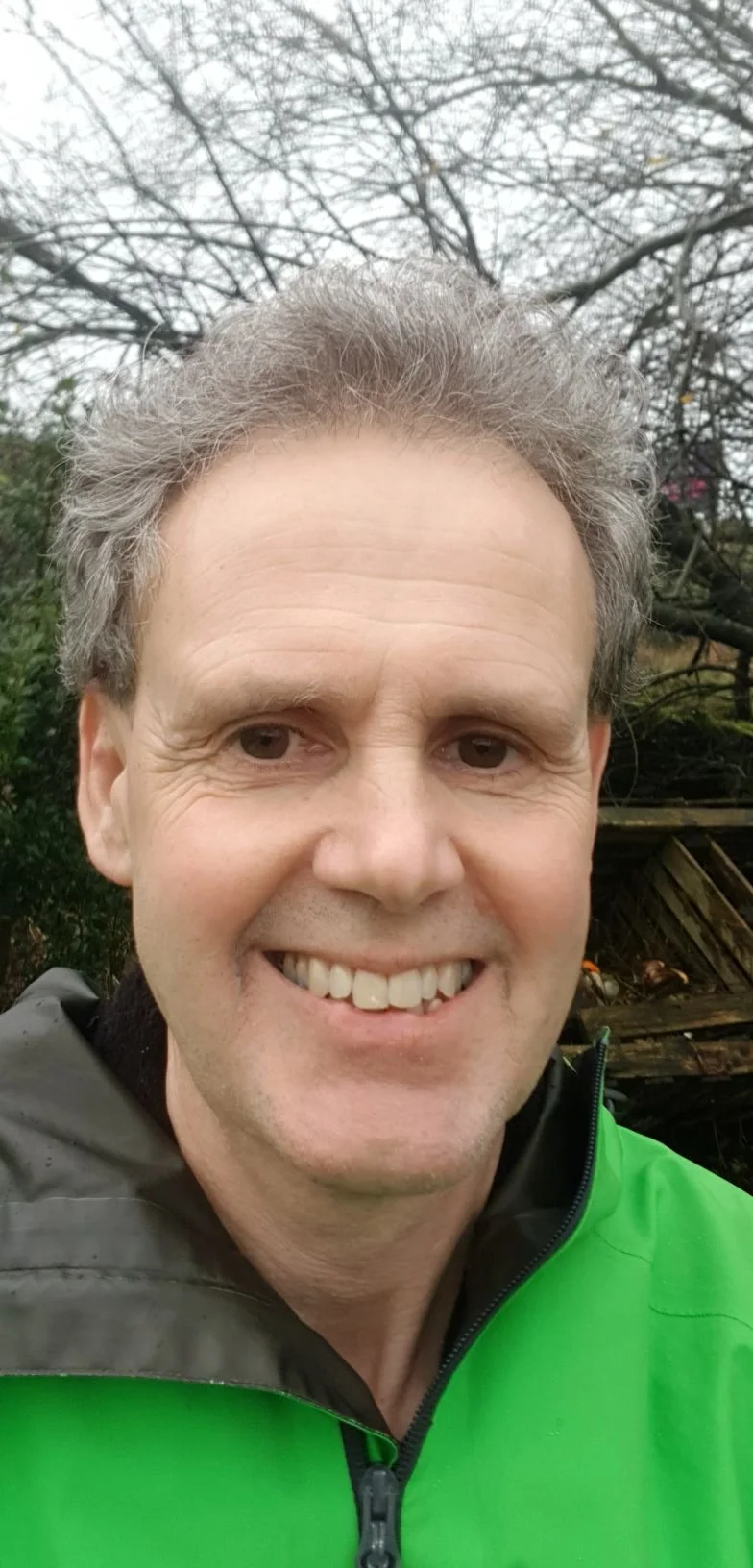 A smiling man outdoors wearing a green jacket with a black collar, with a background of leafless trees and some greenery. Noel Watkins, biodanza yorkshire