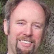 Close-up of a man with light brown hair, a goatee, and a mustache, smiling in front of a blurred outdoor background. Peter Naropa burns biodanza frome