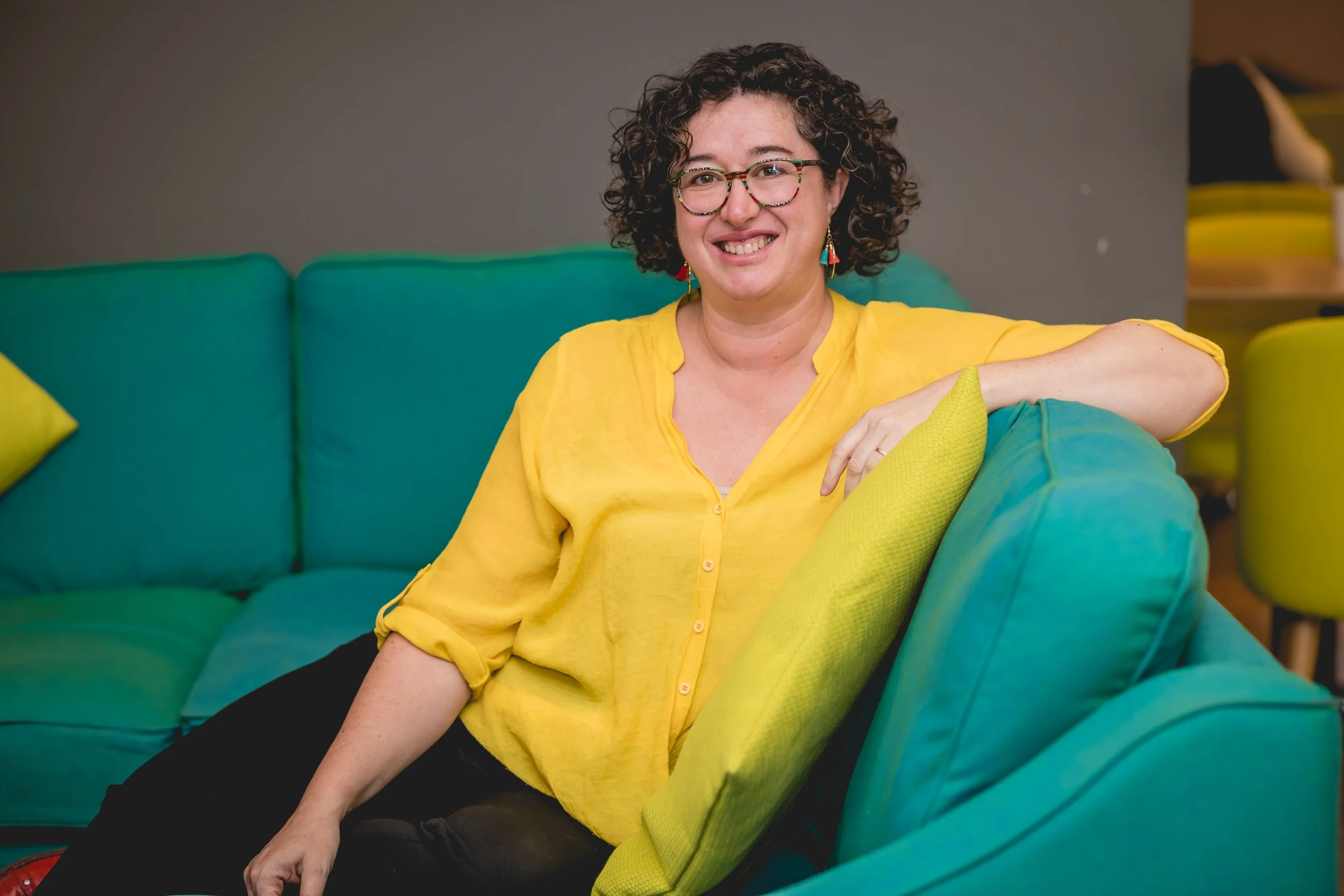 A woman with curly dark hair and glasses, wearing a yellow blouse, sitting on a teal couch with a yellow cushion, smiling at the camera. Peggy Cheyo biodanza milton keynes berkhamsted