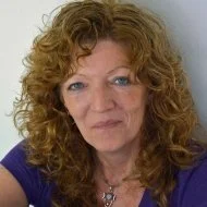 A woman with curly red hair and blue eyes looking at the camera, wearing a navy blue top.