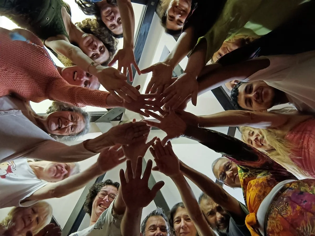 A diverse group of women and men forming a circle with their hands touching in the middle, looking down at the camera with smiles and excitement.