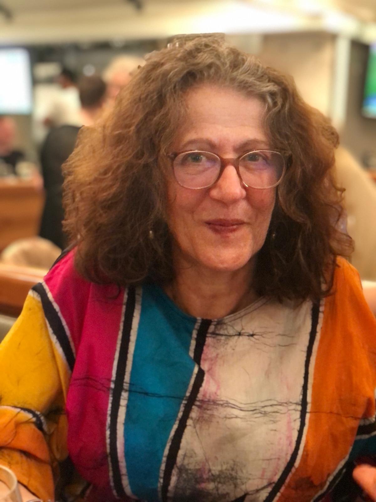A woman with curly hair and glasses, wearing a colorful blouse, seated at a restaurant or cafe. Miriam biodanza london