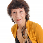 A woman with short, curly dark hair and light skin, wearing a mustard yellow top and a necklace, looking slightly to the side. Antoinette Lorraine, biodanza Cardiff and Bristol. Bristol school of Biodanza