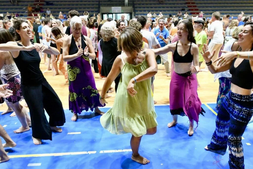 People dancing and enjoying themselves at an indoor event, with a diverse group of women in colorful skirts and tops dancing on a blue floor, surrounded by others in the background.