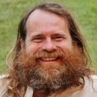A man with long hair and a full beard smiling outdoors on a grassy background. Rupert Meese biodanza