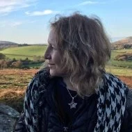 Profile of a woman with curly gray hair wearing a patterned jacket, standing outdoors with a green landscape in the background. olga Bastable biodanza glastonbury, london, rotingean