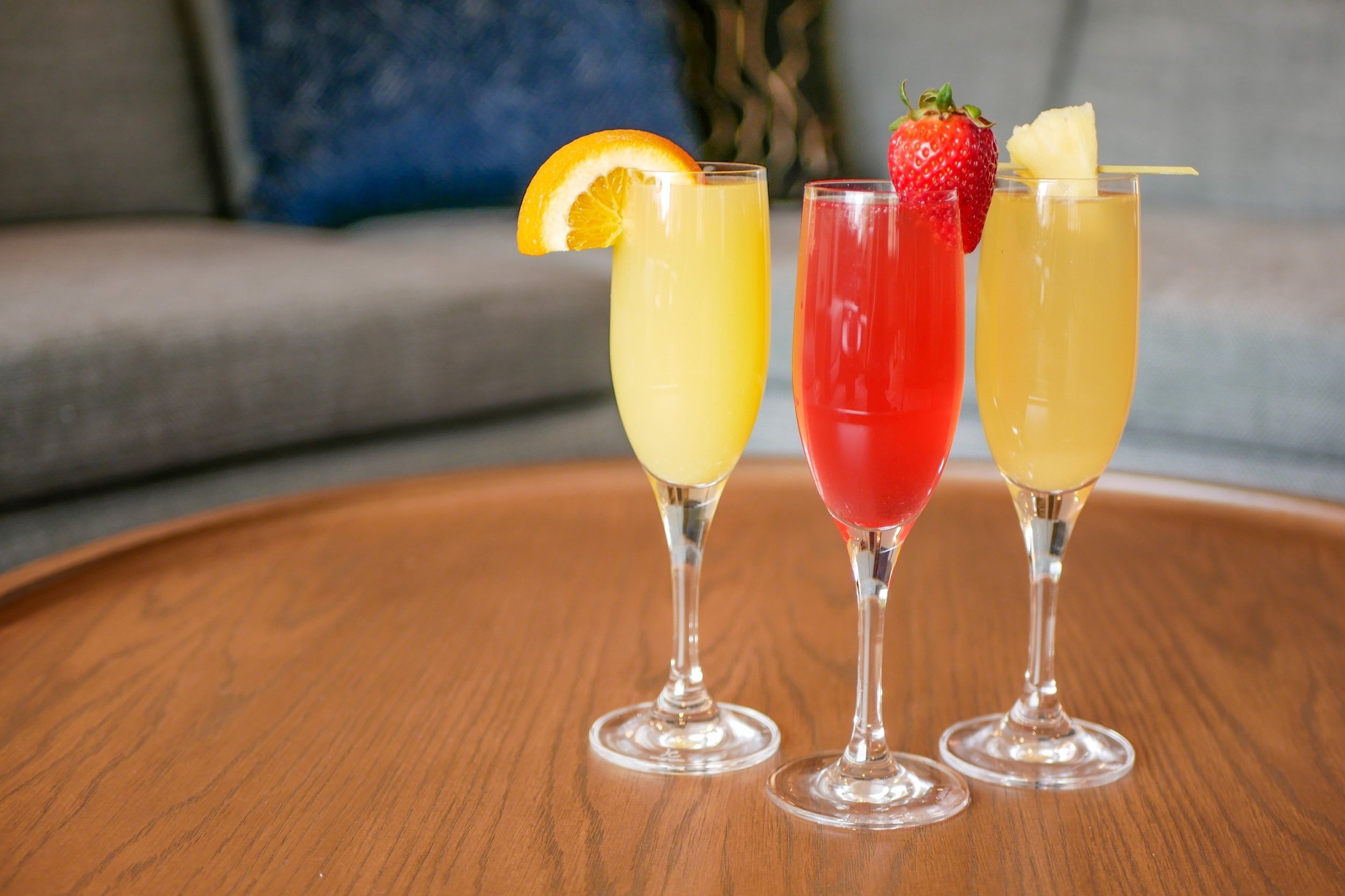 Three colorful cocktail drinks in fluted glasses on a wooden table, garnished with fruit slices and berries.