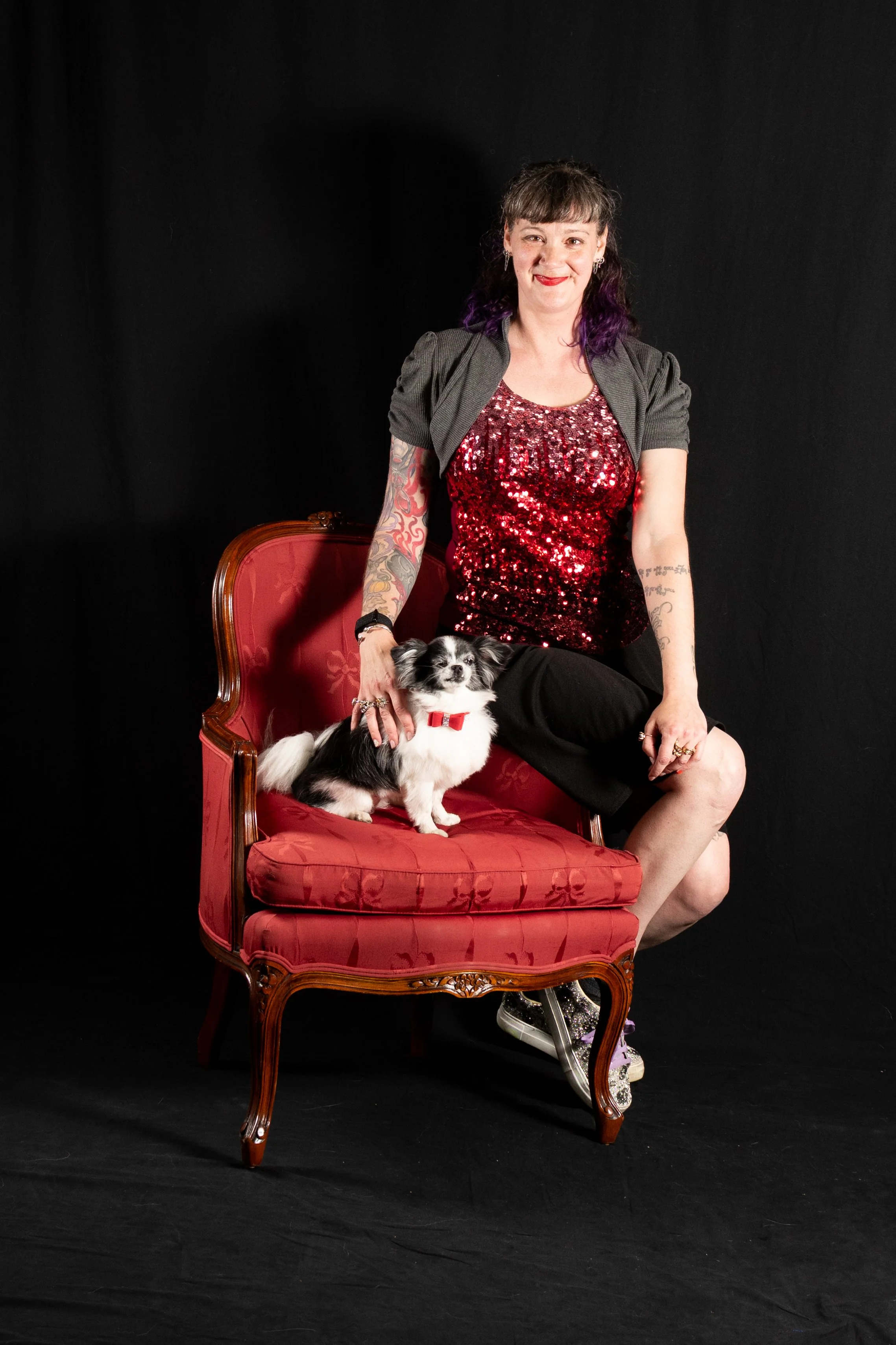 A woman with tattoos on her arms, wearing a red sequined top and a dark jacket, sitting on an ornate, red velvet vintage chair with wooden details, holding a small black and white dog with a red collar. The background is black.