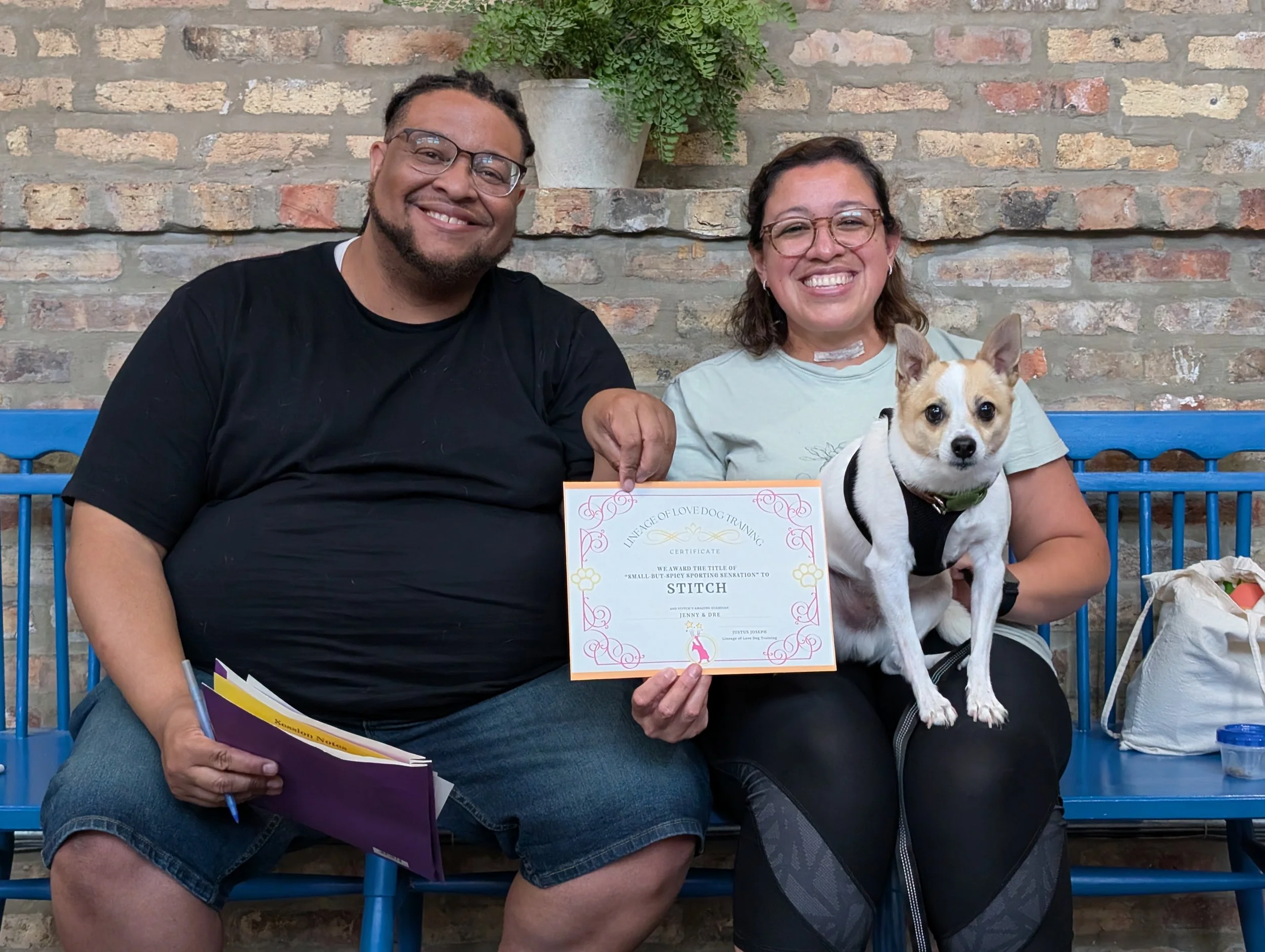 A smiling man with glasses and beard, holding a certificate, sitting next to a woman with glasses, smiling, holding a dog and a purple folder, in front of a brick wall with a potted plant. They appear to have just completed a dog training class.