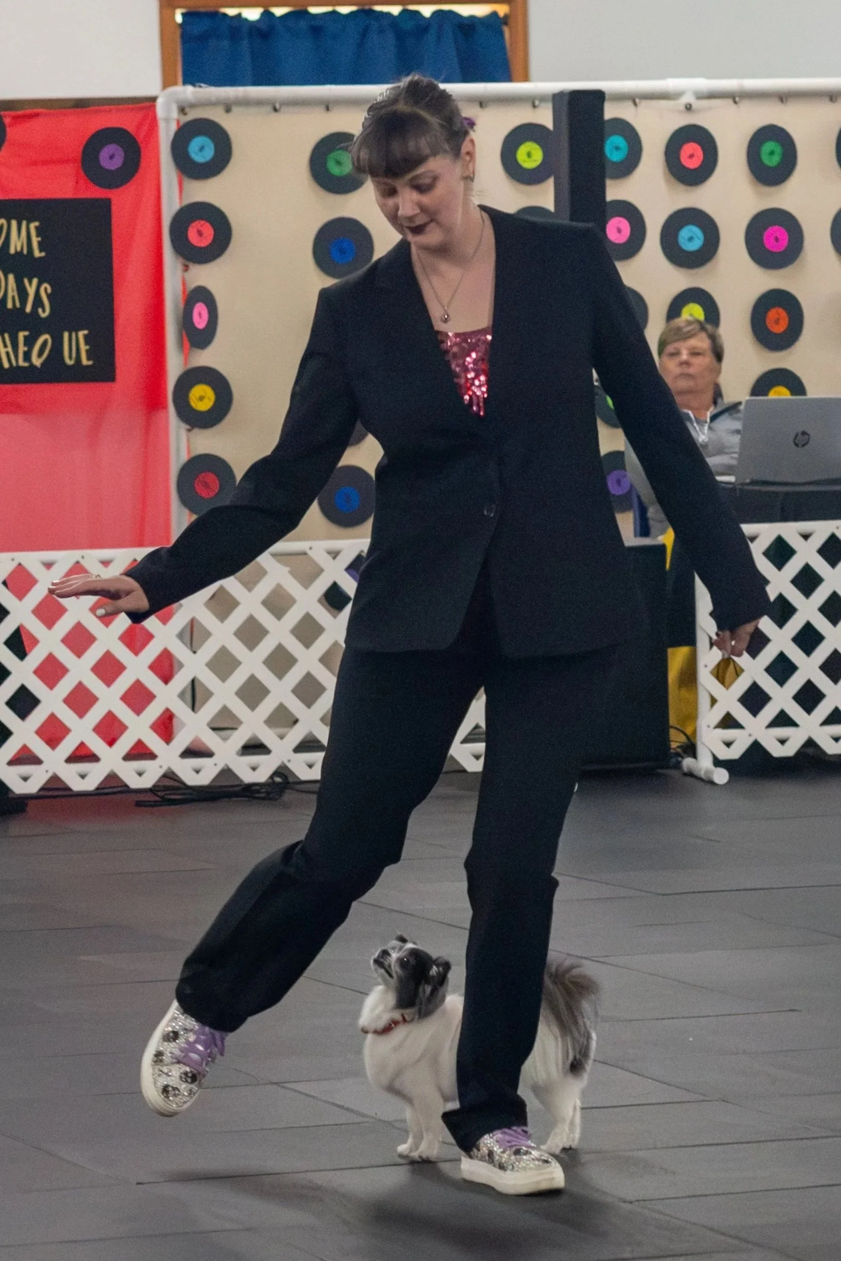 A woman in a black blazer and sequined top is performing a dance of some kind with a small dog on a leash. They are dancing on a stage with a backdrop decorated with colored records and a sign that reads 'WELCOME DAYS AHEAD UE.'